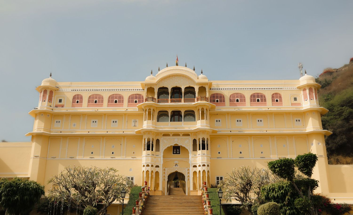 Grand yellow Rajasthani palace with arched balconies, ornate facade, and manicured gardens under a hazy sky in Jaipur, India.
