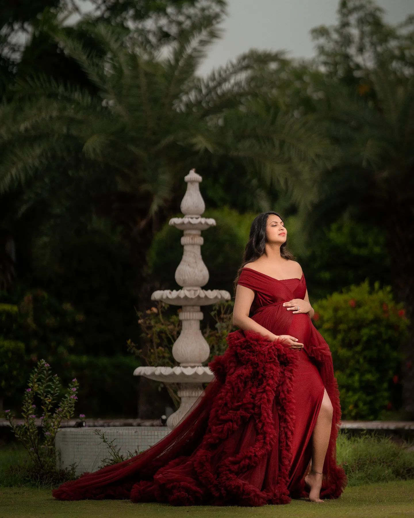 An elegant maternity portrait in a red gown next to a garden fountain.