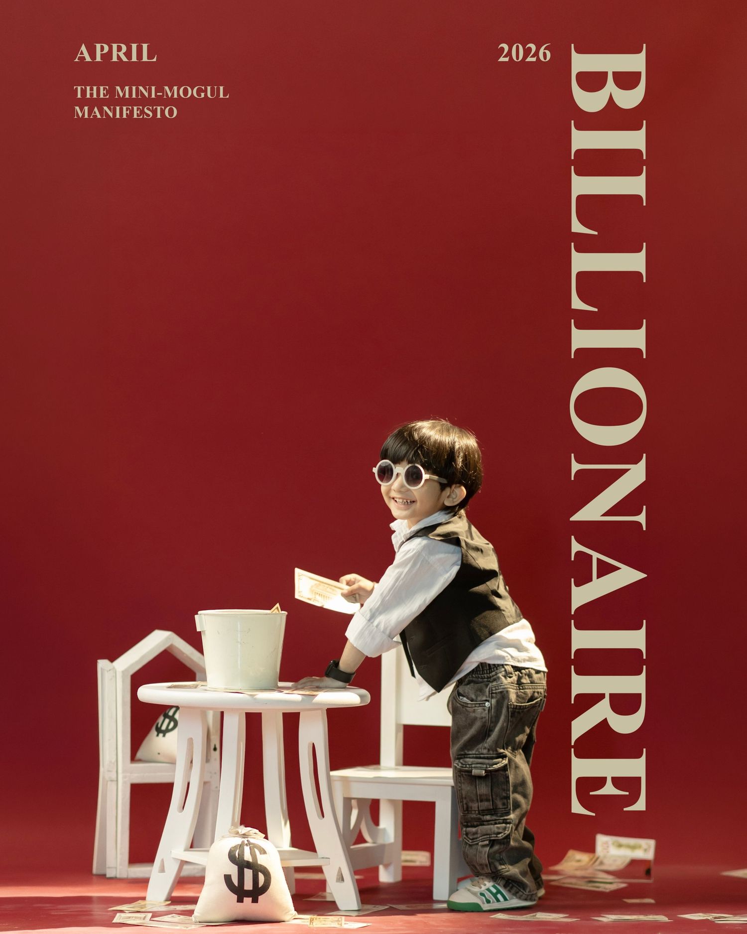 A young child dressed as a mini mogul poses with money bags and a white desk on a red background for a Billionaire magazine cover.