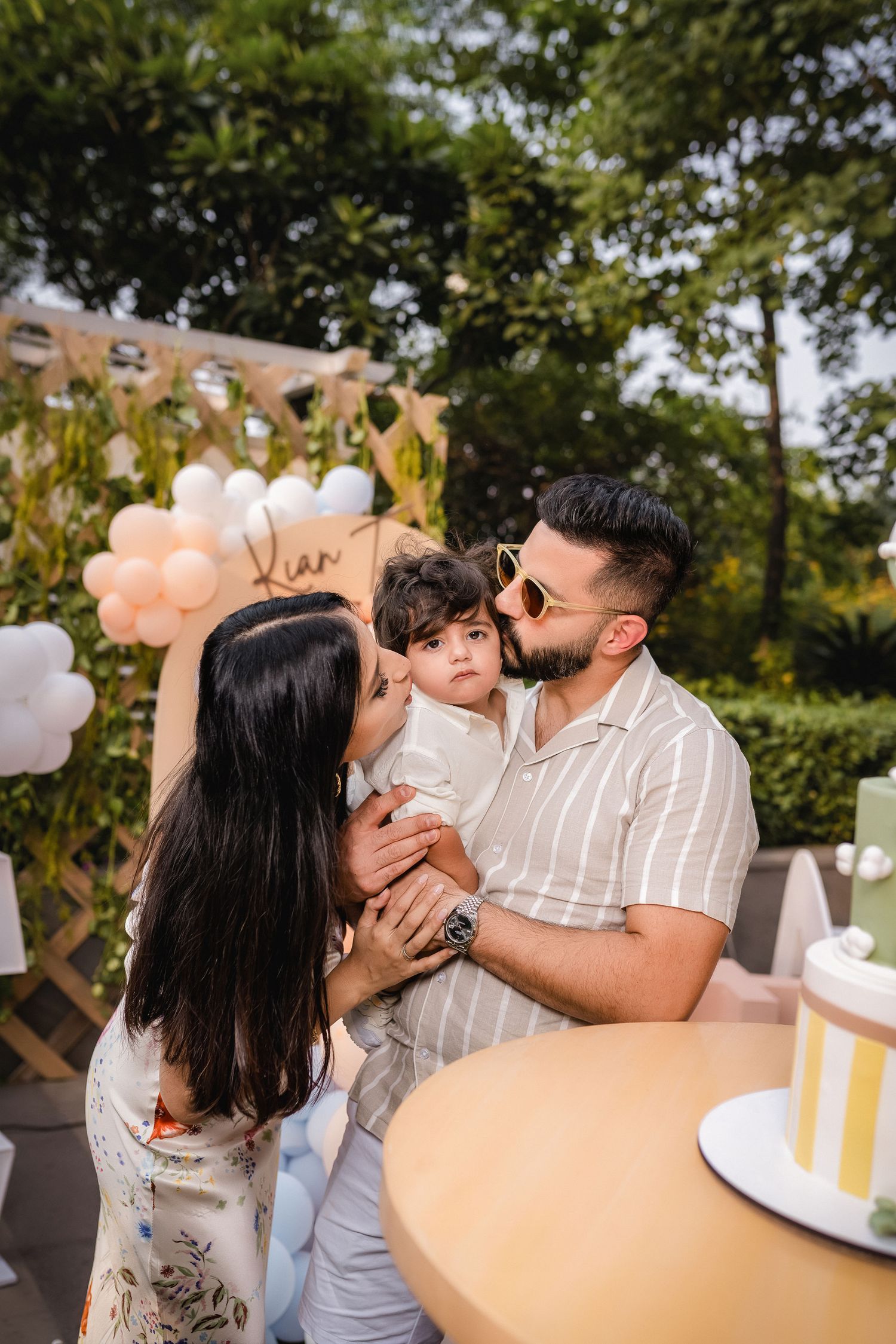 Parents kissing their toddler at an outdoor birthday party with balloon decorations and a cake.