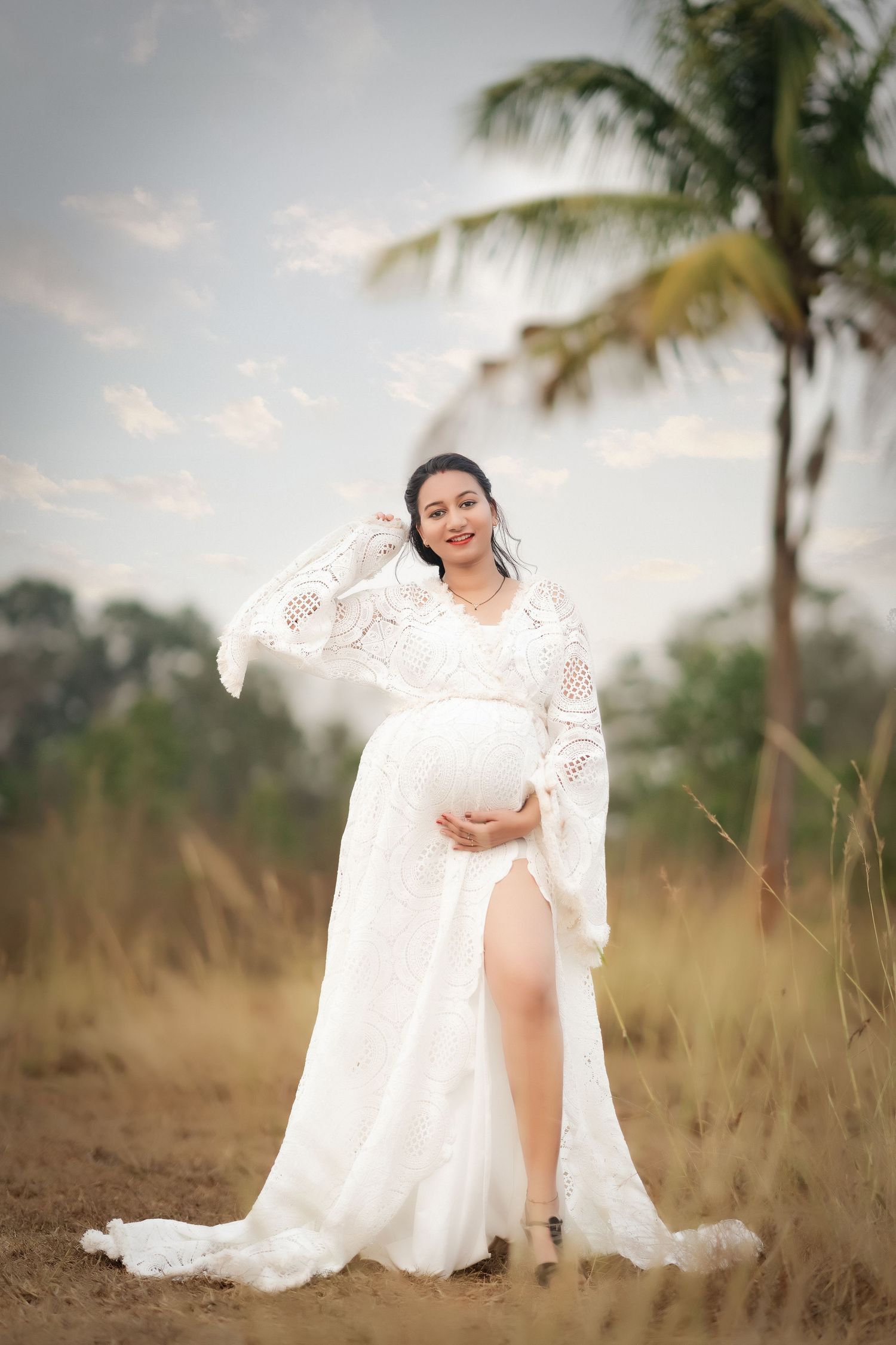 Pregnant woman in white lace maternity gown poses outdoors near palm trees in a serene nature setting.