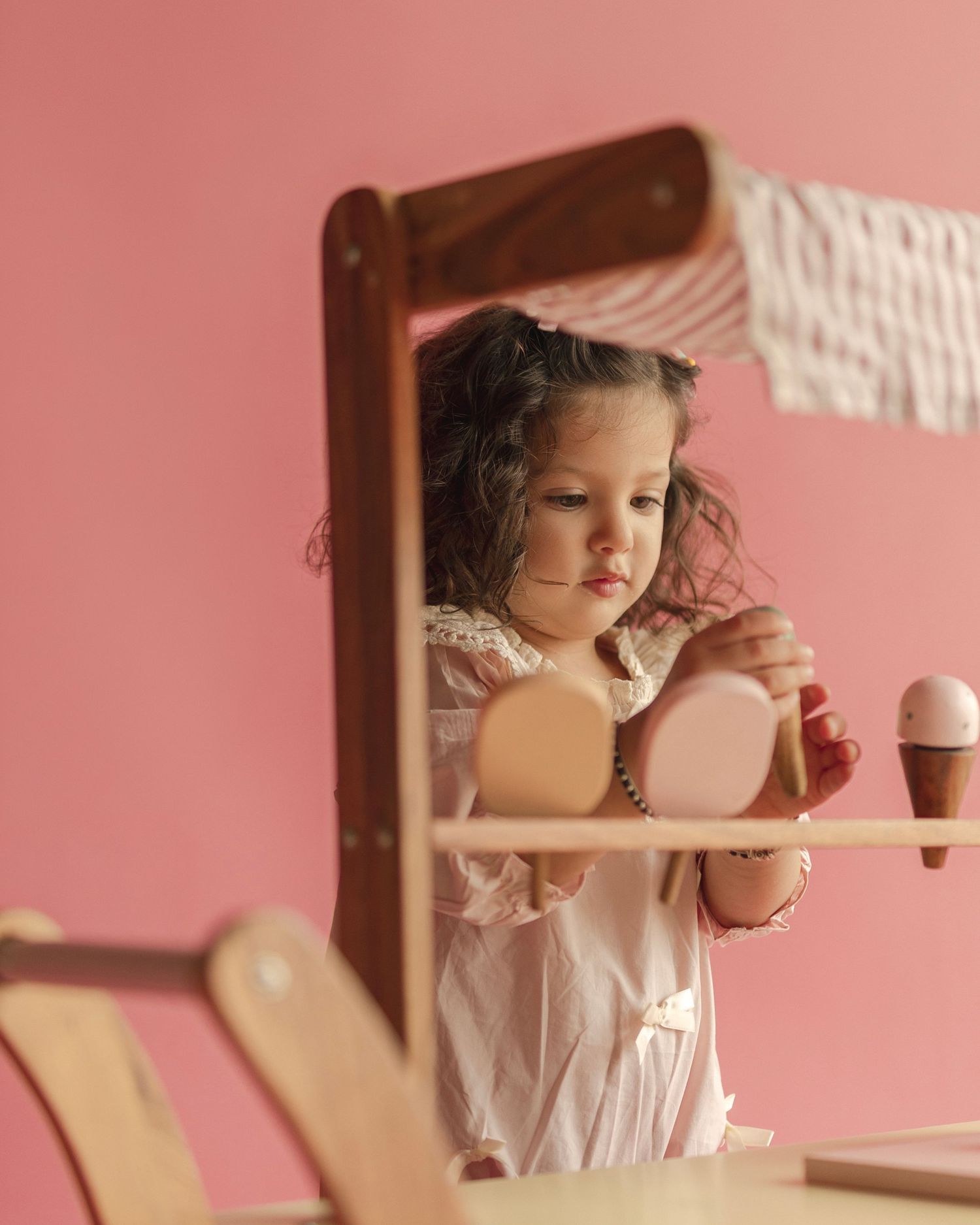 A young girl with curly hair plays with wooden toy ice cream set against a pink background.