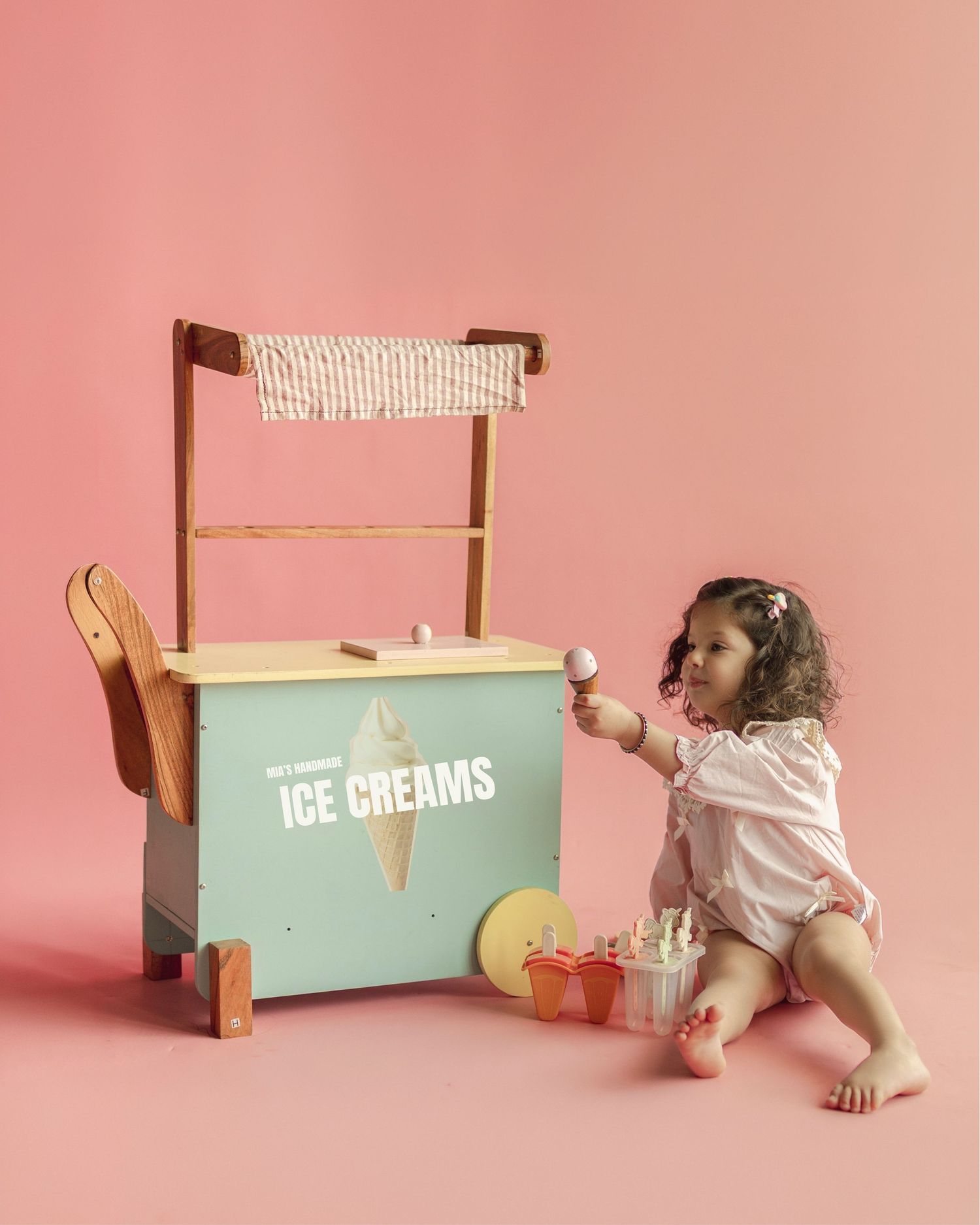 Toddler girl in pink romper playing with a mint green wooden ice cream cart toy on a pink background.