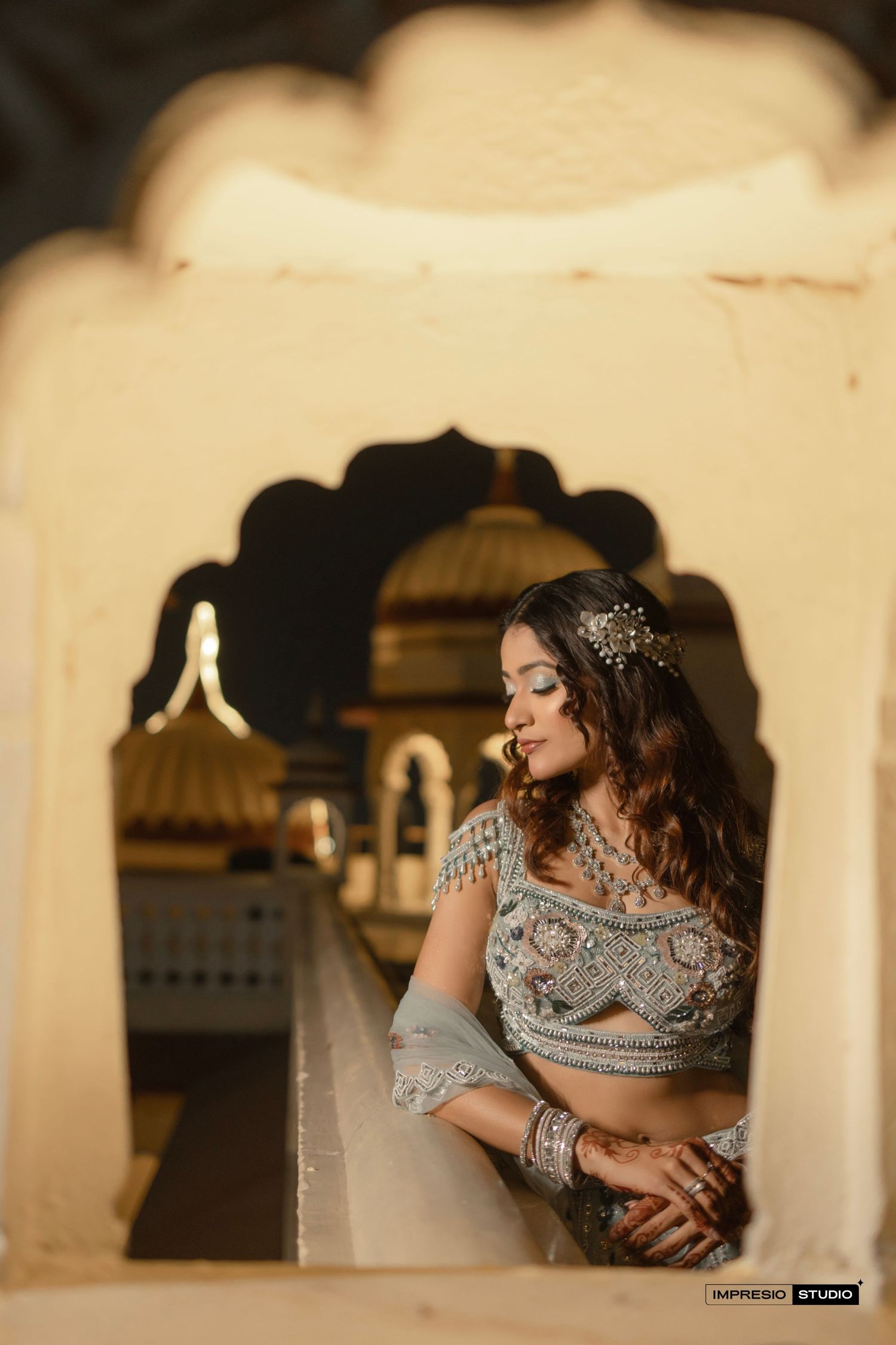 A bride in silver lehenga poses gracefully under ornate Rajasthani arch, adorned with jewelry and hair accessories.
