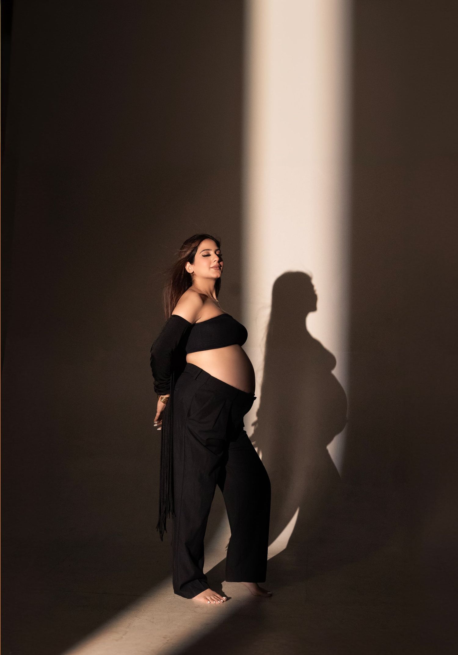 Pregnant woman in black off-shoulder top and pants poses gracefully in dramatic light, casting a beautiful silhouette shadow.