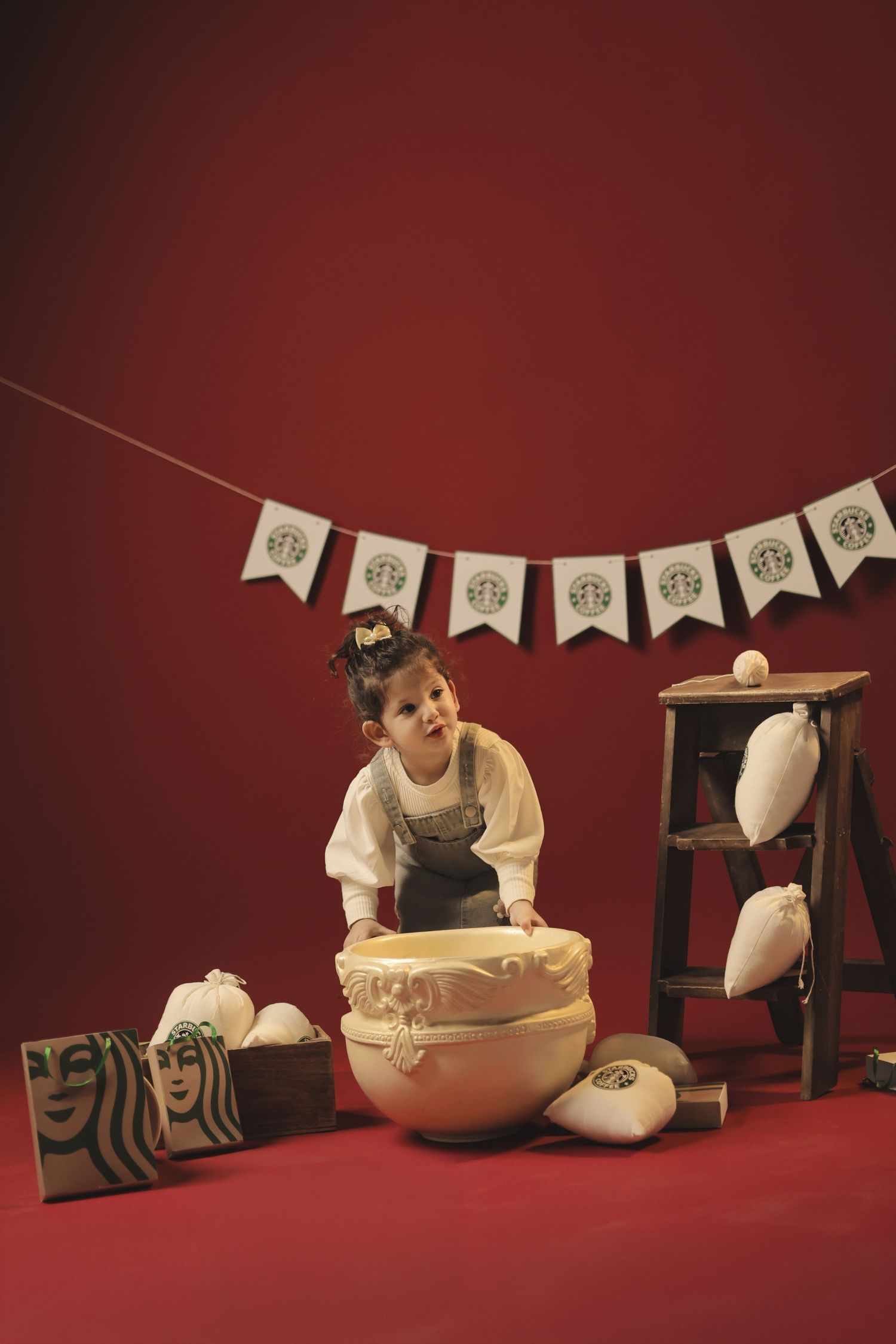 Child sits with large Starbucks bowl surrounded by branded gifts and bunting on red background.