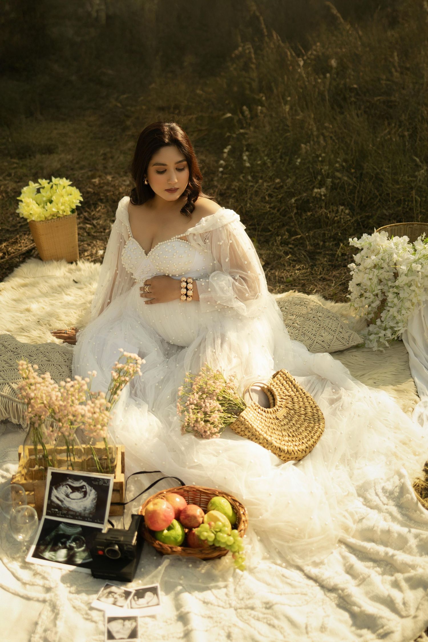 Maternity picnic scene with white dress, flowers, fruit basket and ultrasound photos on a blanket.