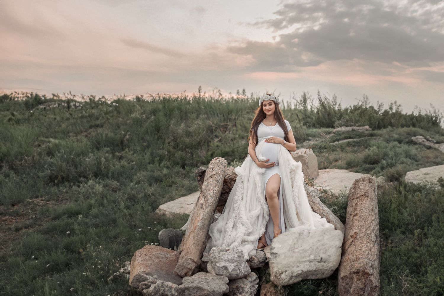 A sunset maternity portrait of someone wearing white dress seated on natural rock formation.