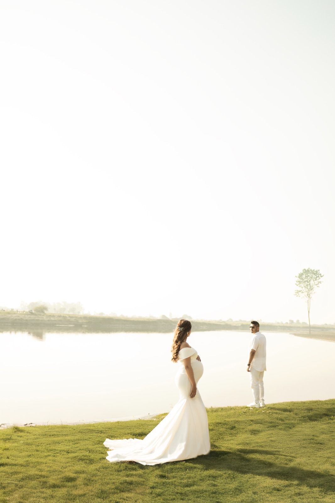 A couple in wedding attire poses on a grassy hill overlooking a serene lake during golden hour.