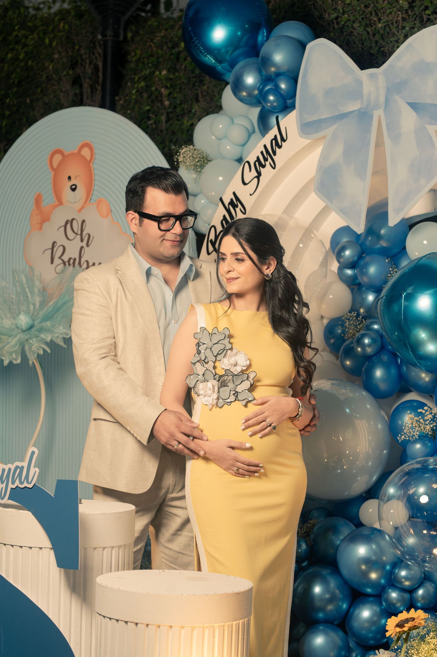 Couple at a blue and white baby shower with balloon arch, pregnant woman in yellow dress, man in beige suit, Baby Sayal sign.