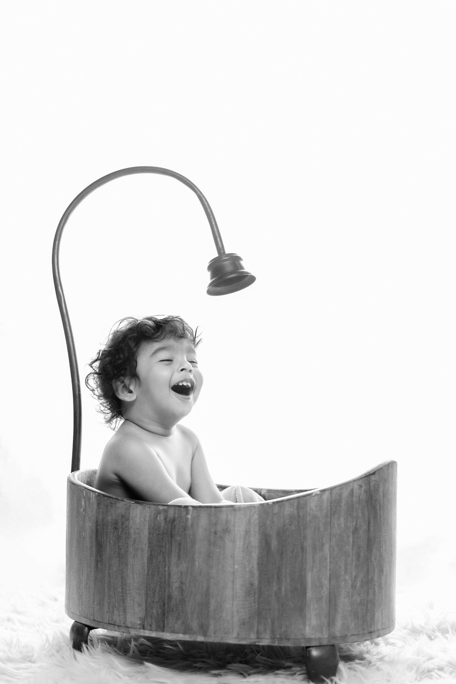 Black and white photo of a laughing baby sitting in a vintage wooden bathtub with a shower head.