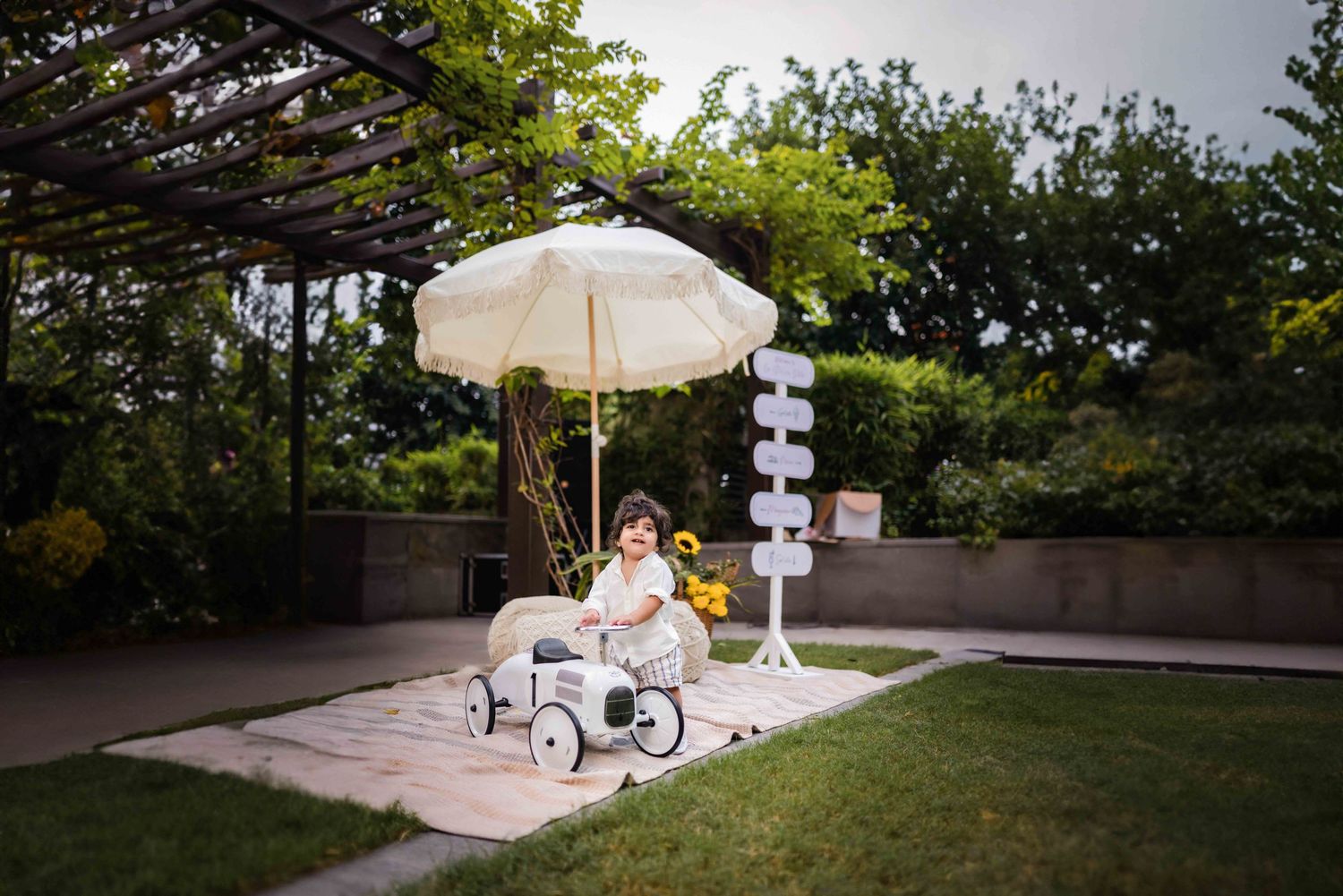 Child sitting in a vintage toy car under a cream umbrella in a lush garden setting with a decorative sign post.