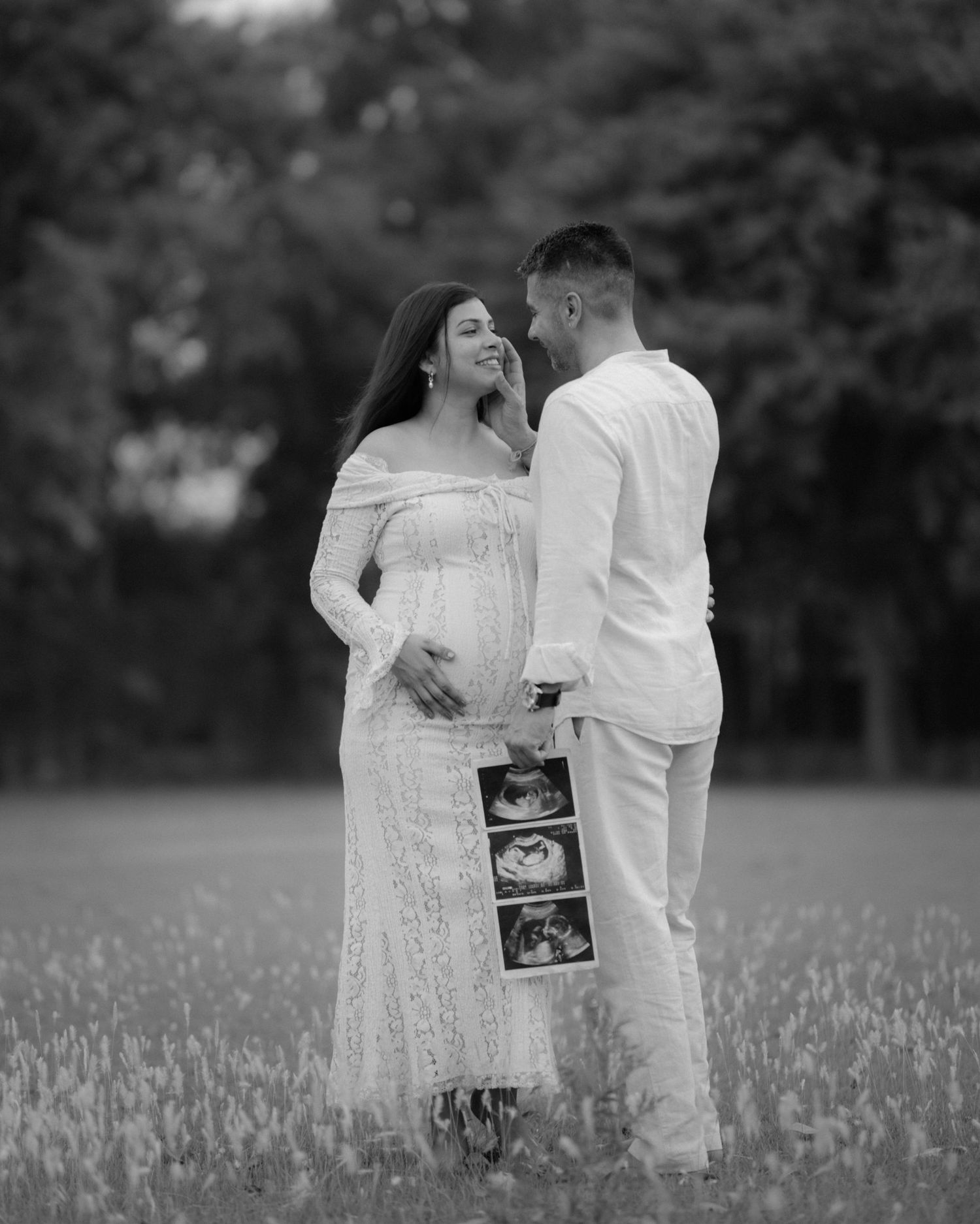 Black and white maternity photo of pregnant couple in field, woman in lace gown, man holding ultrasound images.
