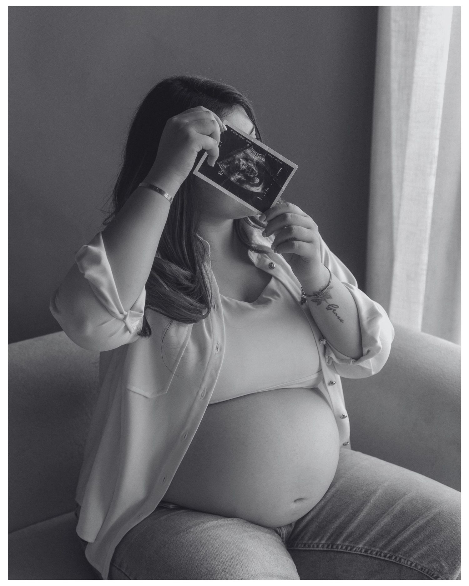 Black and white photo of a pregnant woman holding an ultrasound image over her face while sitting on a couch.