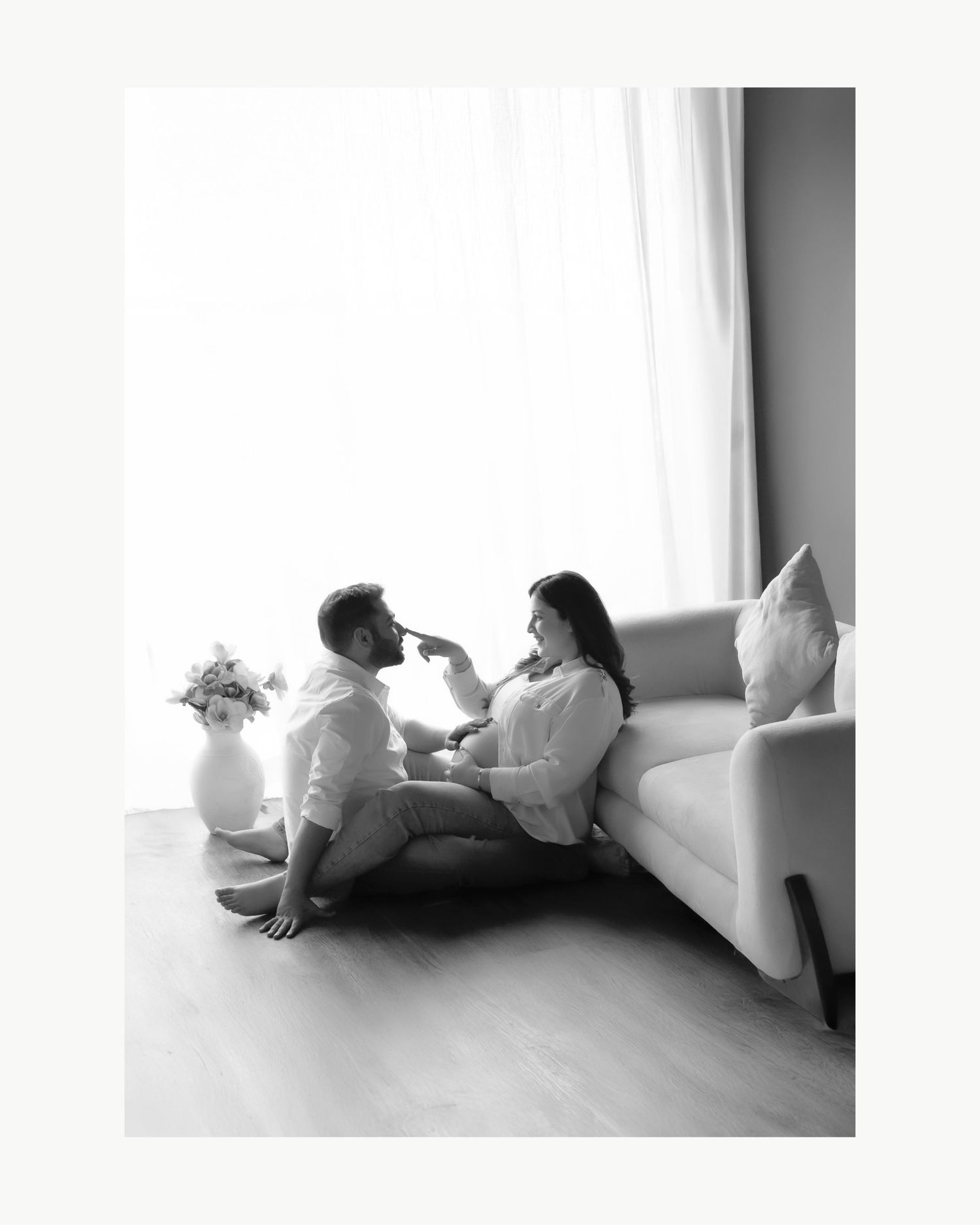 Black and white photo of a couple sitting on the floor by a sofa, sharing a tender moment near a bright window.