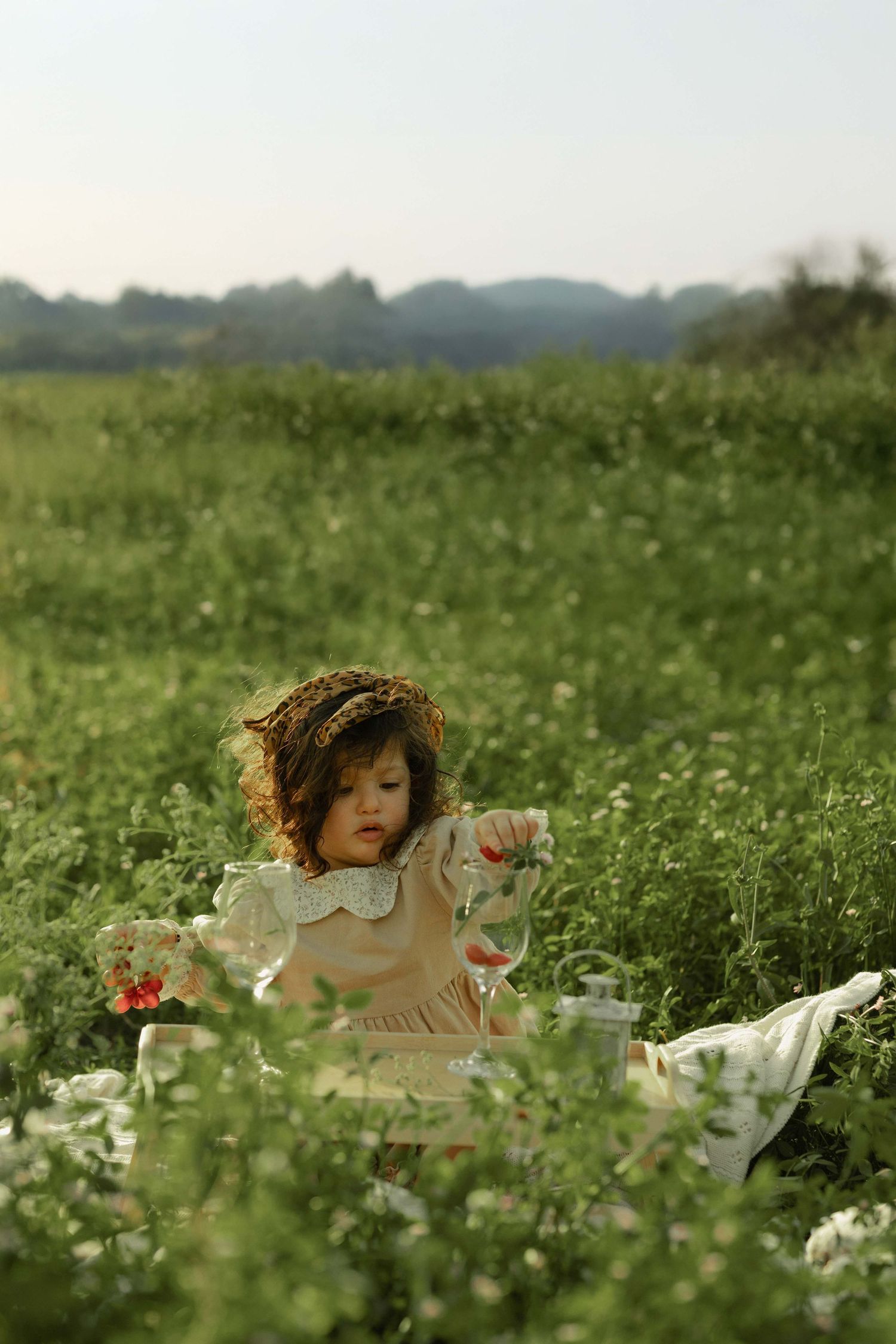 Young girl in floral dress and straw hat sitting in a lush green meadow with wildflowers on a sunny day.