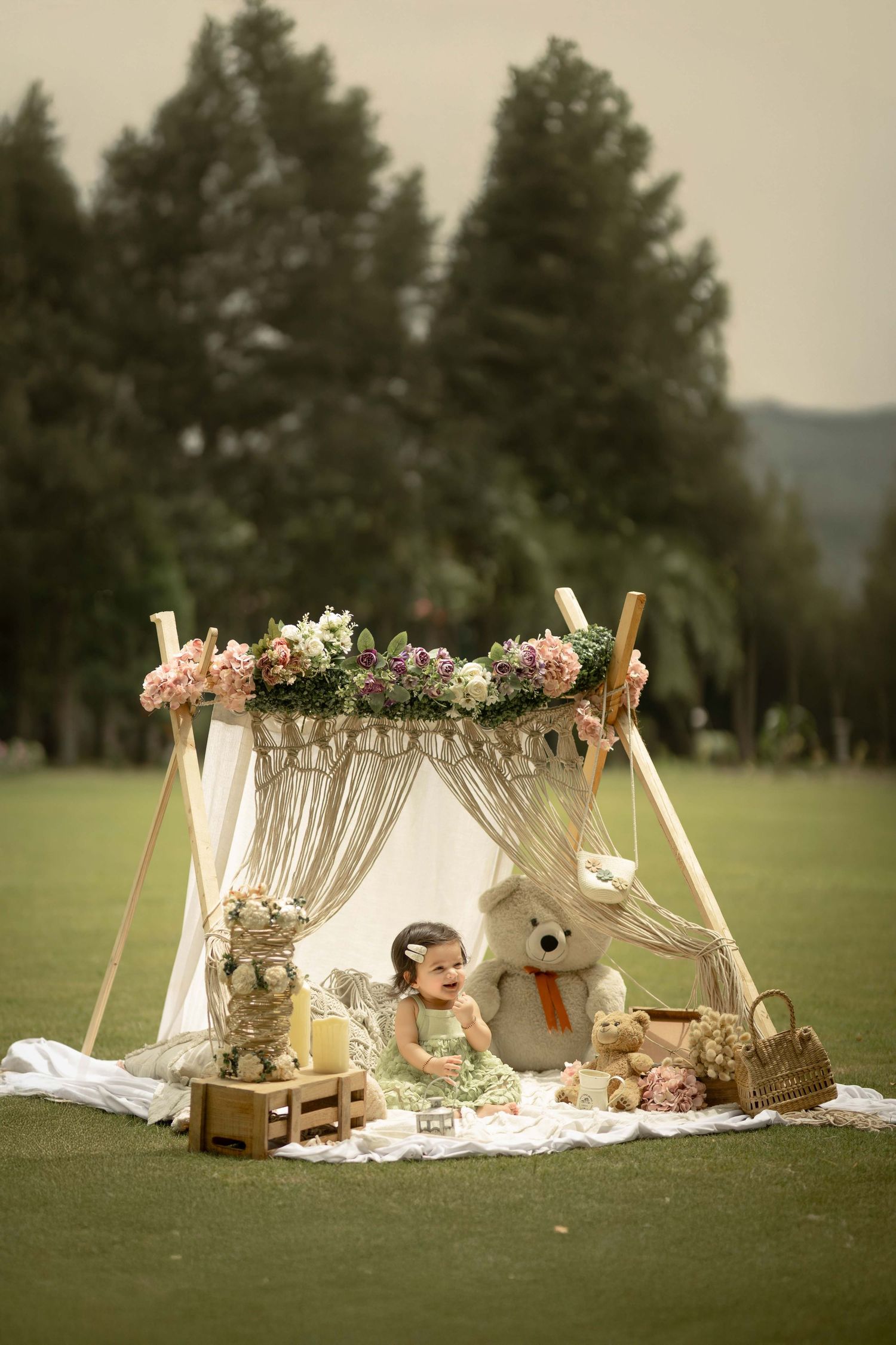 Baby girl in green dress laughing beside teddy bear under floral boho tent on grass field.