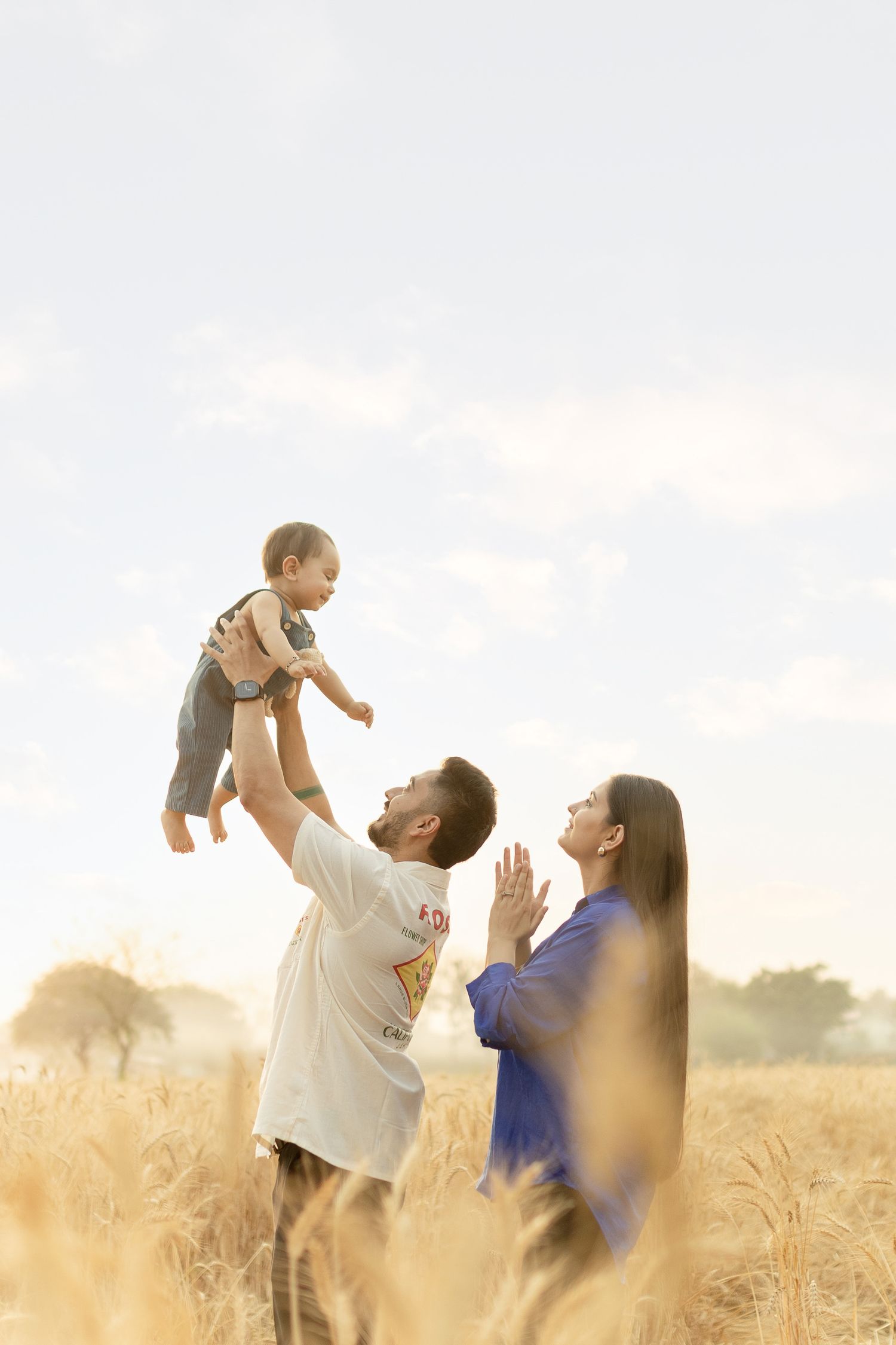 A joyful family in a golden wheat field, father lifting baby high while mother watches with hands clasped in delight.