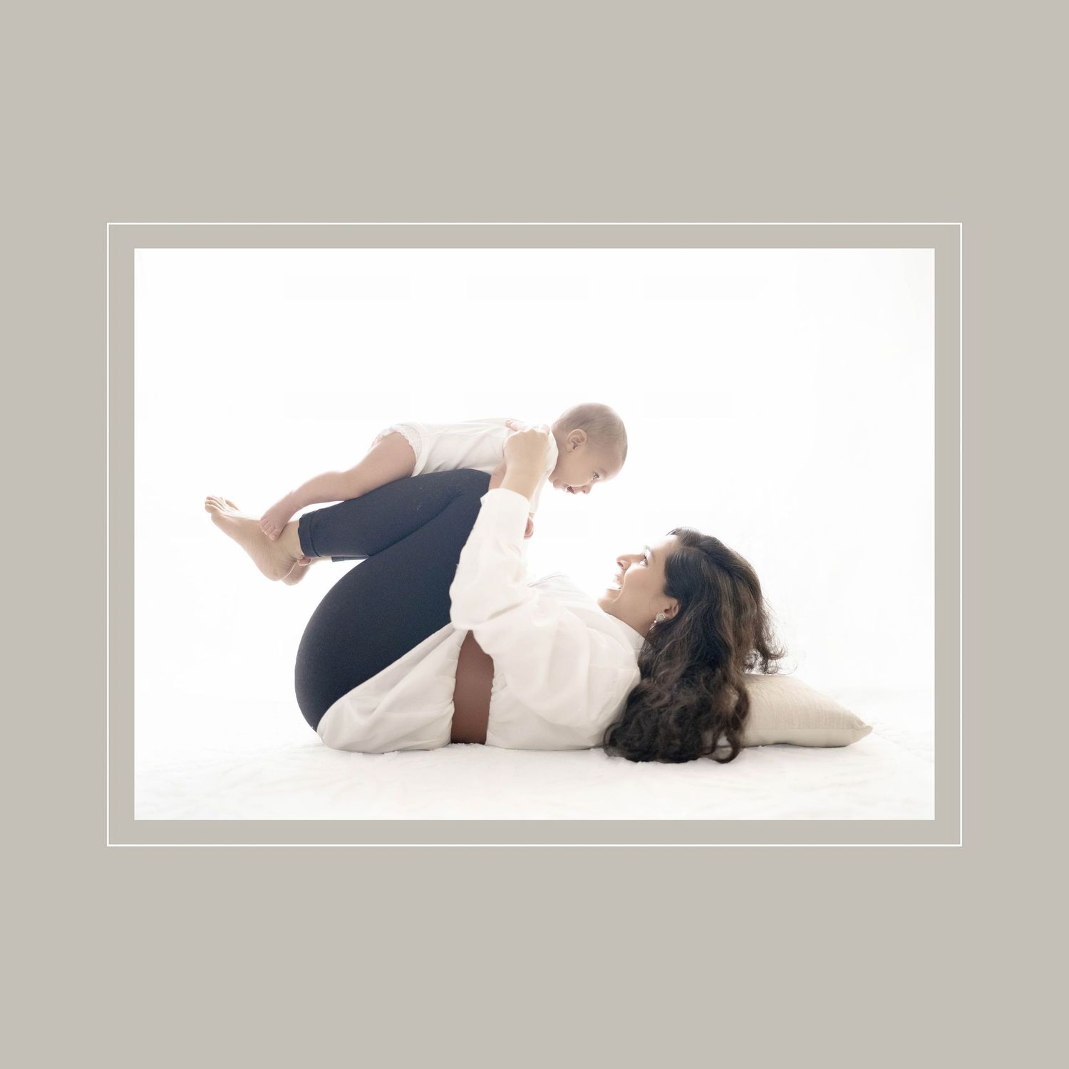 A mother lies on her back lifting her baby in the air during a tender white-background photo session.