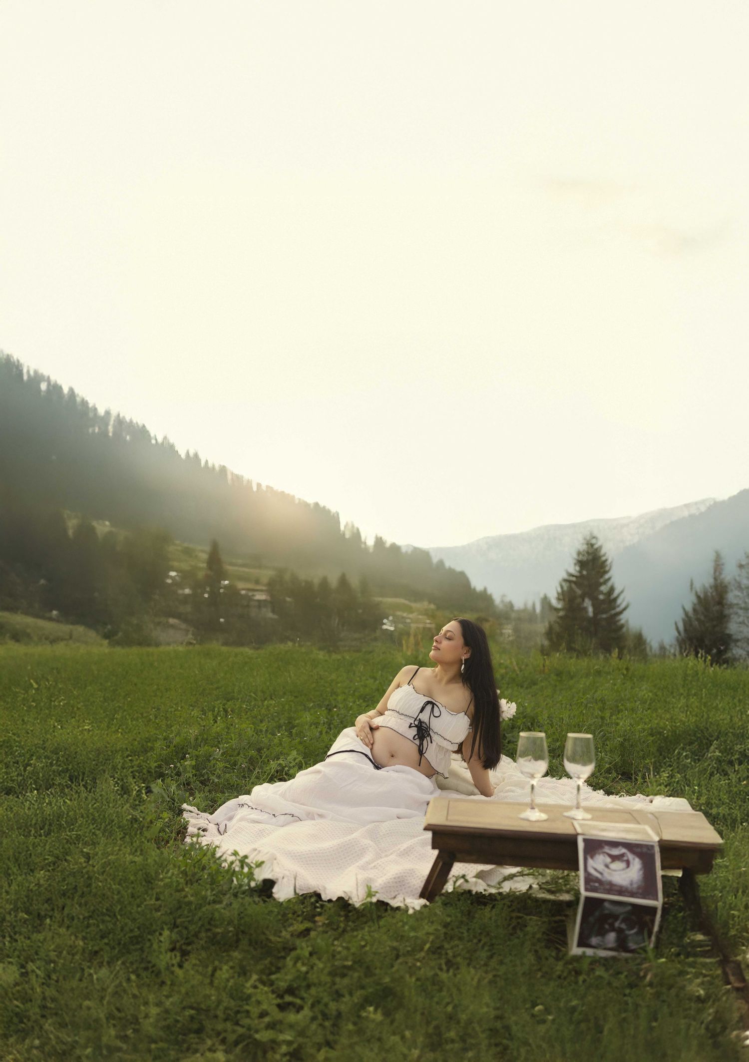 Pregnant woman in white dress relaxing at a scenic mountain meadow picnic with wine glasses at sunset.