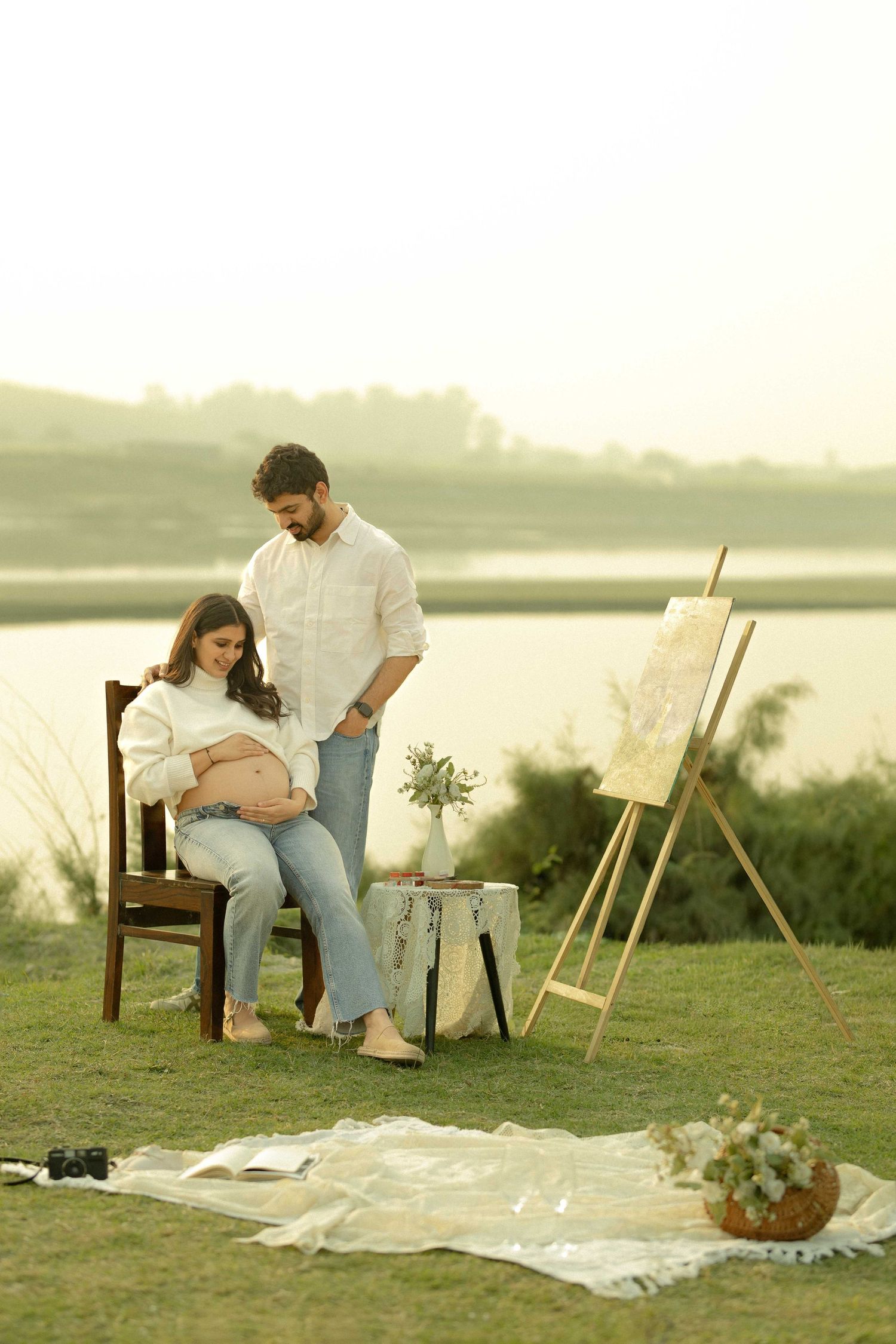 Pregnant woman sitting on chair with partner outdoors near lake, maternity photoshoot with easel and picnic setup at sunset.