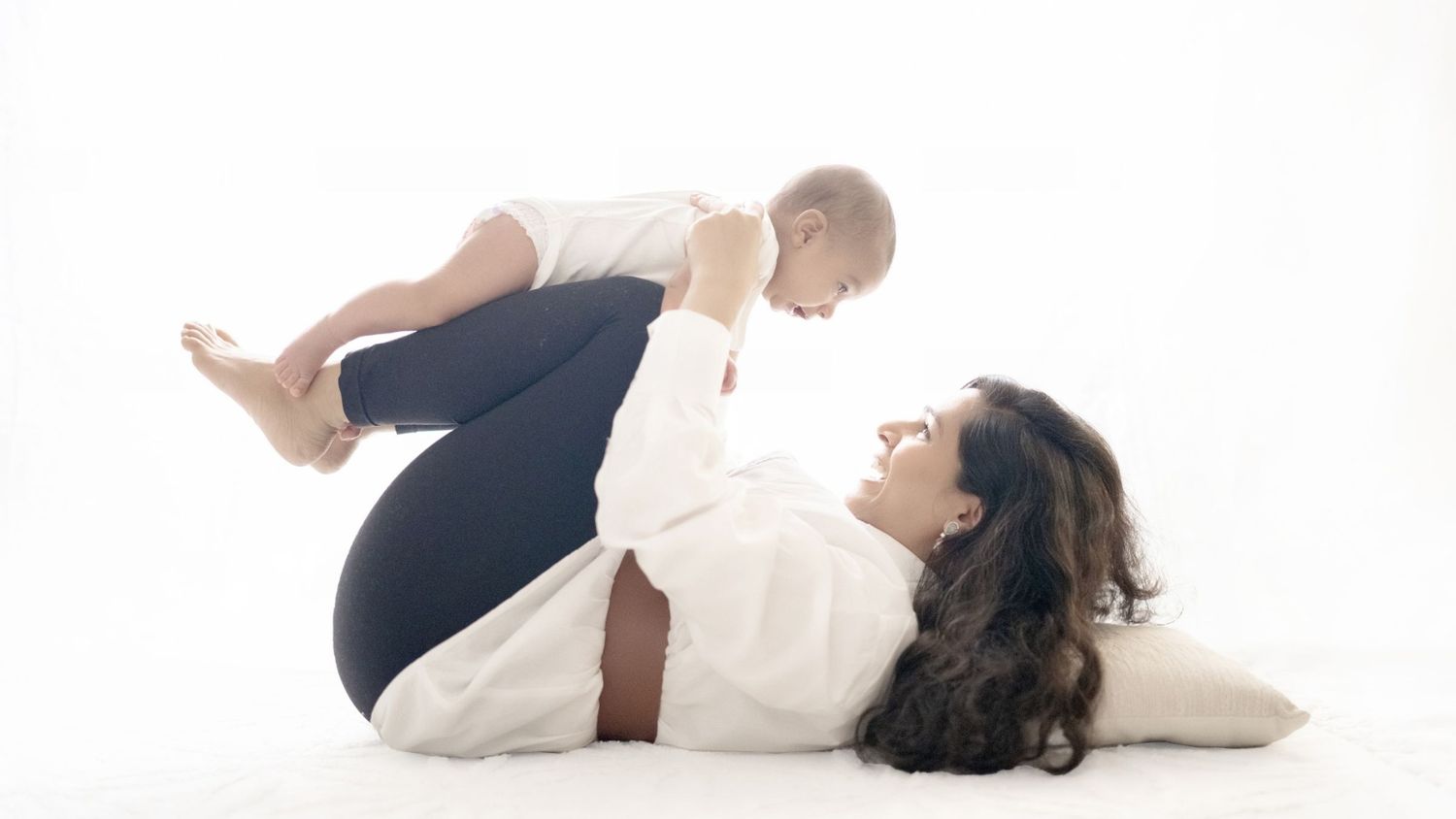 Mother lying on white floor doing baby yoga, lifting smiling infant above her in a bright studio setting.