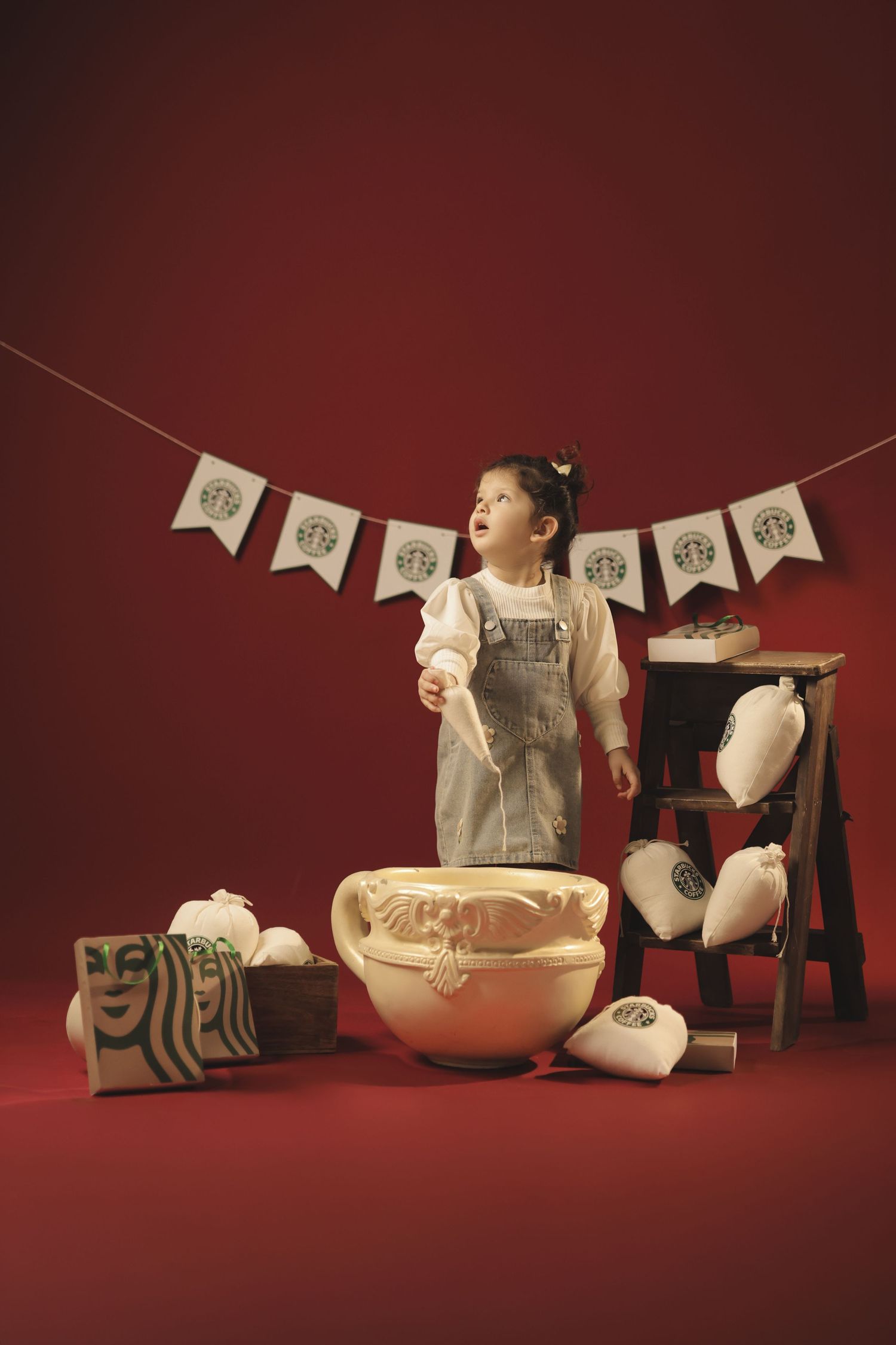 Young girl standing among Starbucks-themed decorations, plush toys, and a large bowl against a red background.