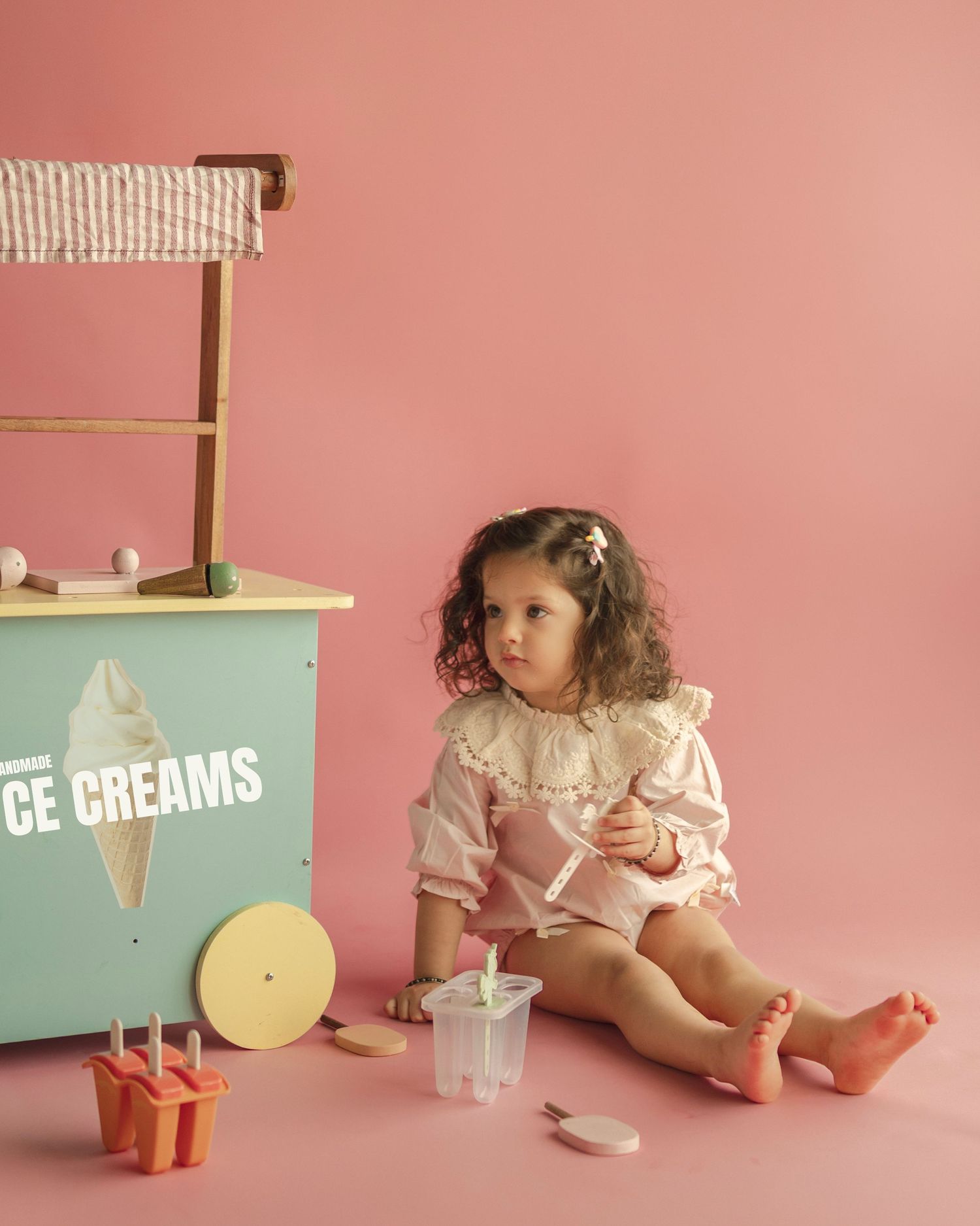 Toddler girl in pink ruffled romper sits beside a mint green wooden ice cream cart on a pink background.