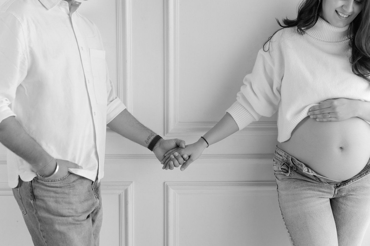 Black and white photo of a pregnant couple holding hands, showing baby bump in jeans and white sweater.