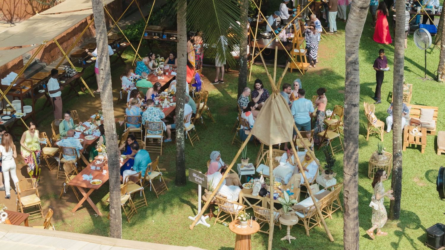 Aerial view of an outdoor tropical dining event with guests seated at wooden tables under palm trees.