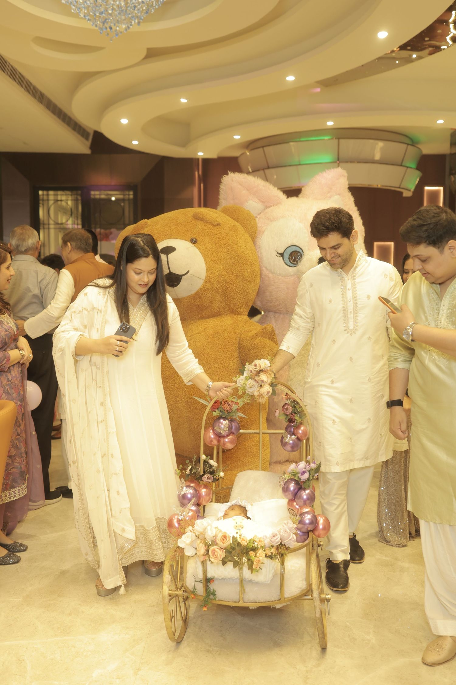 Family in traditional white Indian attire gathers around a flower-decorated baby carriage with a giant teddy bear at a celebration.