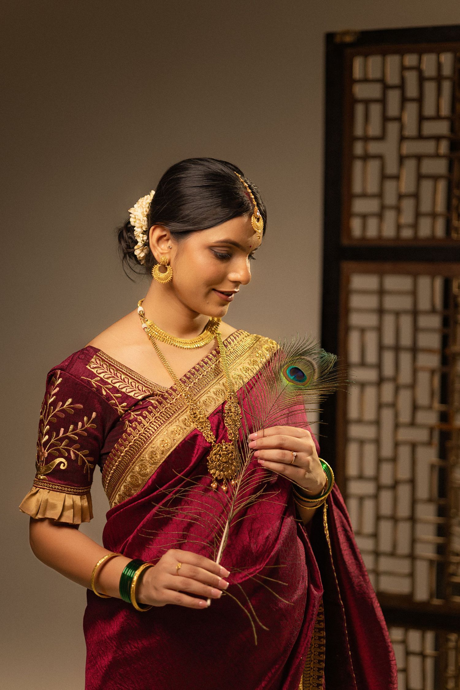 Woman in burgundy and gold traditional Indian saree poses elegantly with a peacock feather in studio lighting.