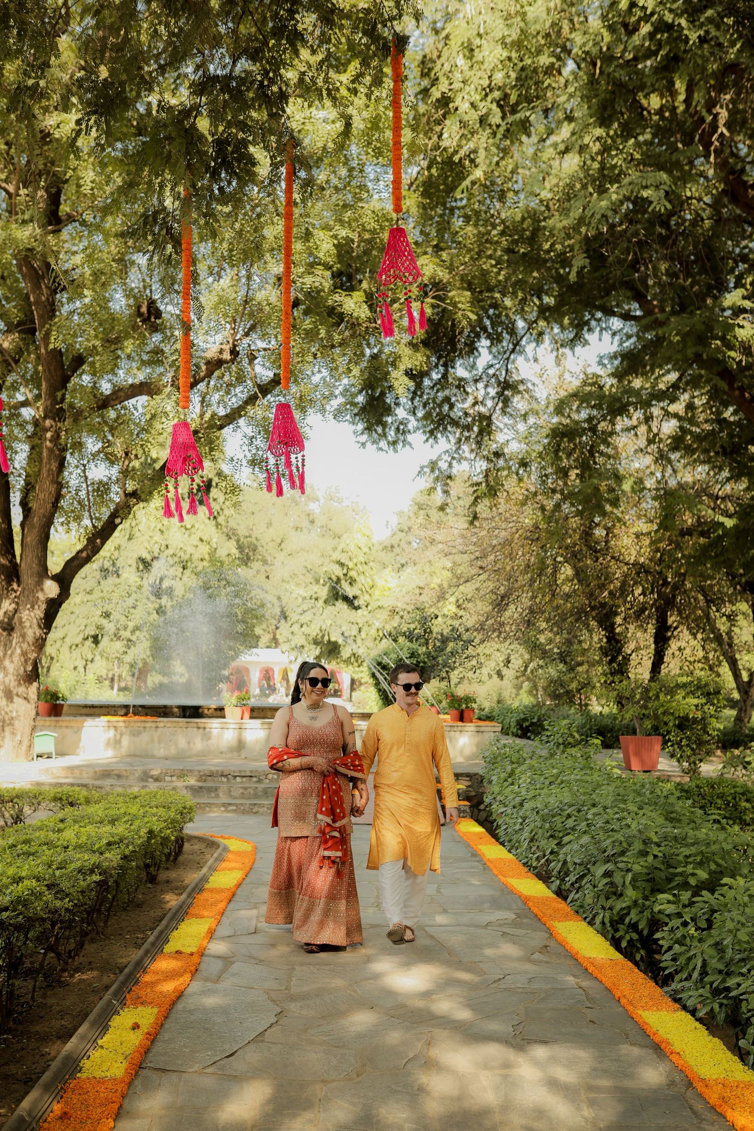 Couple in traditional Indian attire walking on a decorated garden path with colorful tassels hanging from trees.