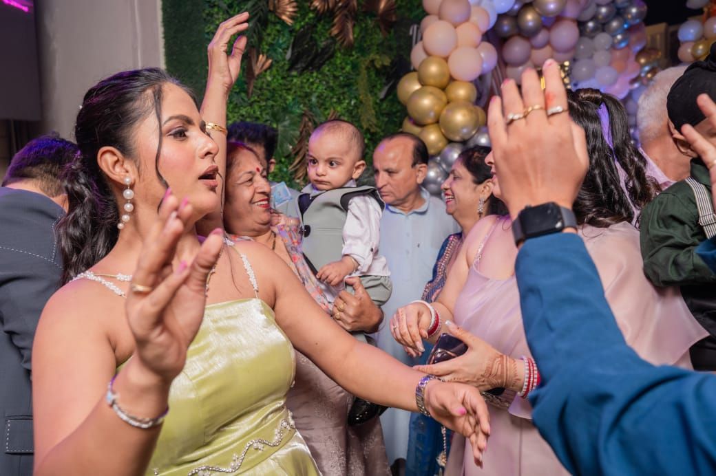 Family celebrating at a first birthday party, dancing and laughing with baby and colorful balloon backdrop.