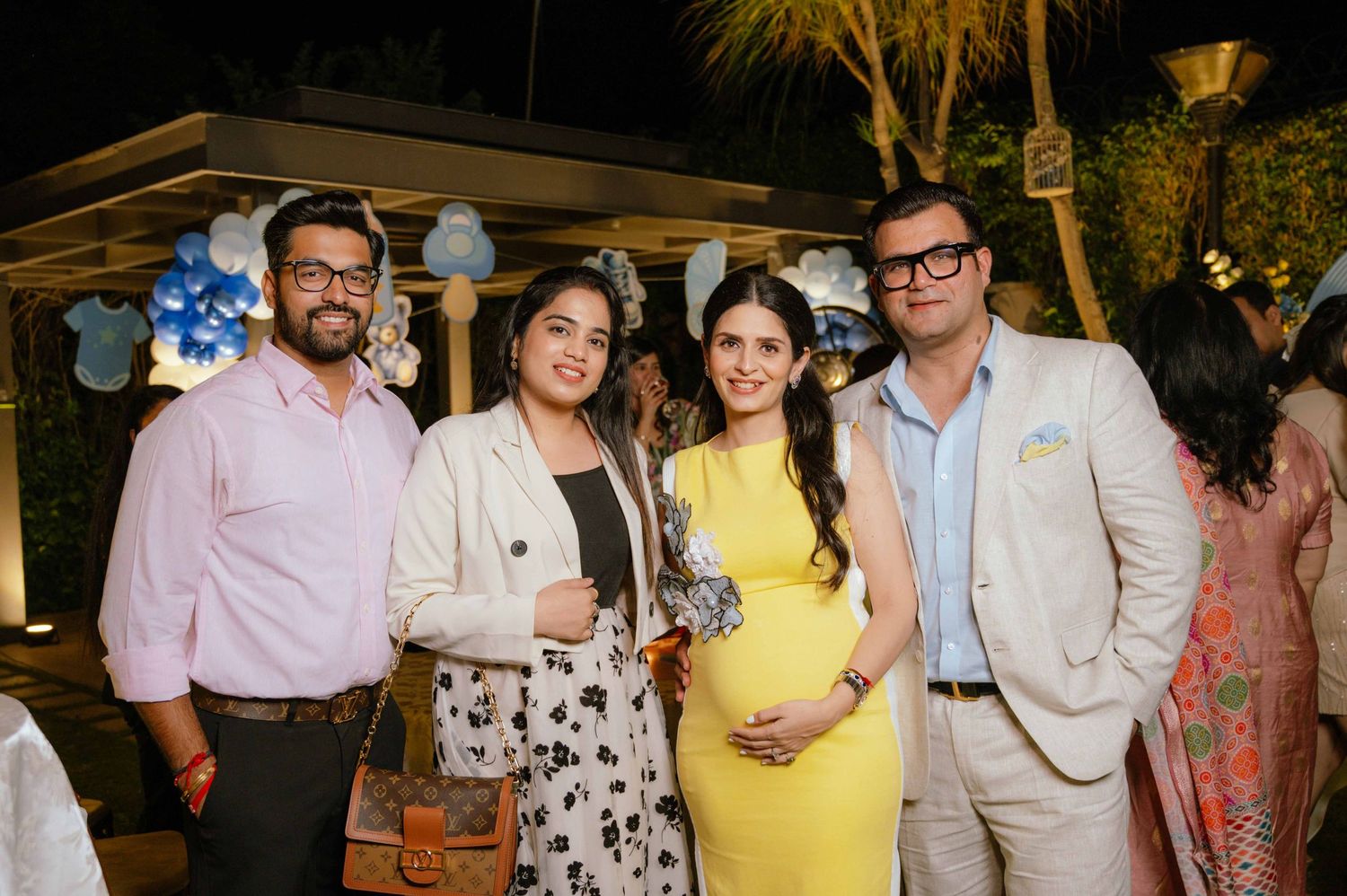 Four friends pose together at a baby shower celebration, with a pregnant woman in yellow dress at center.