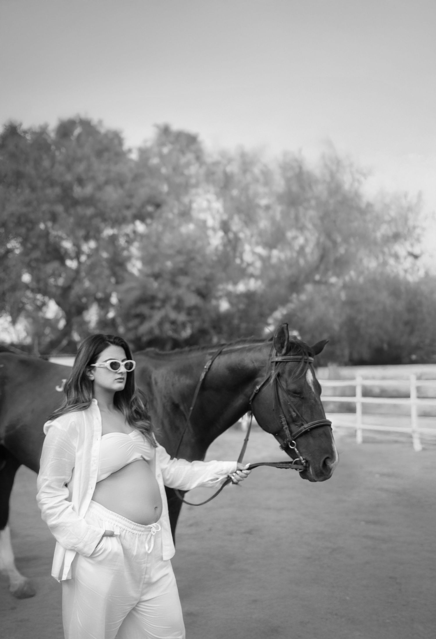 A person in a white blazer poses with a dark horse in an outdoor maternity photoshoot surrounded by white fencing.