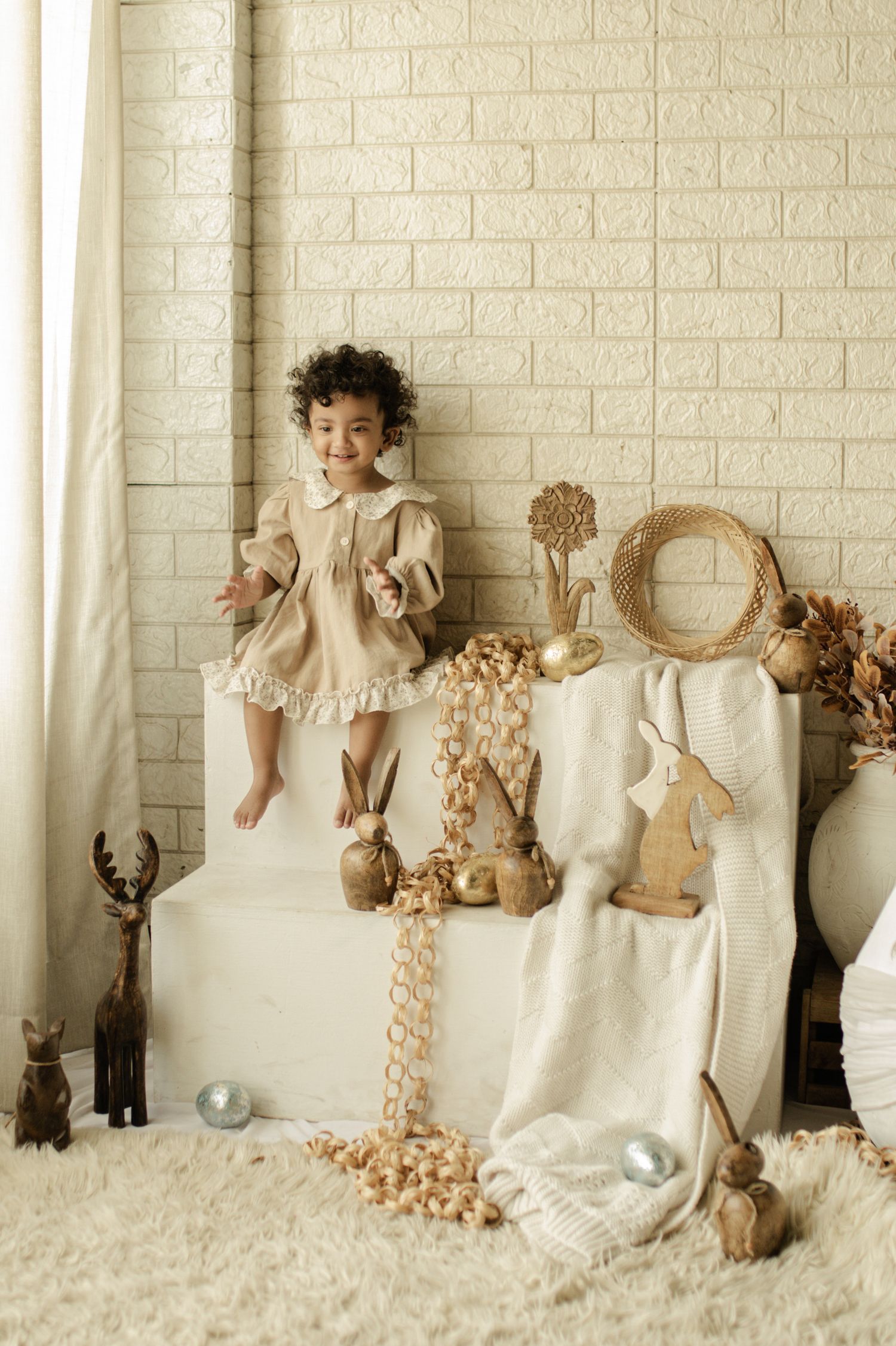 Young girl in beige dress sits on white ottoman surrounded by Easter decorations, wooden bunnies, and wicker accents.