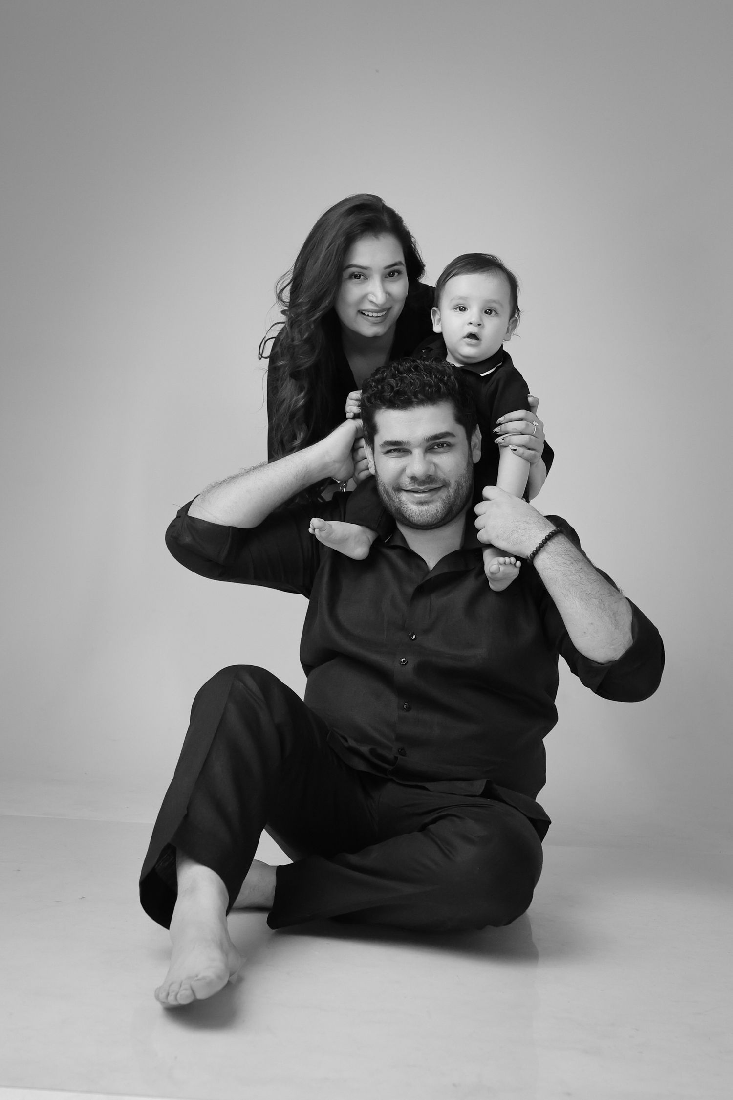 Black and white family portrait of parents and baby, father seated with child on shoulders, mother standing behind.