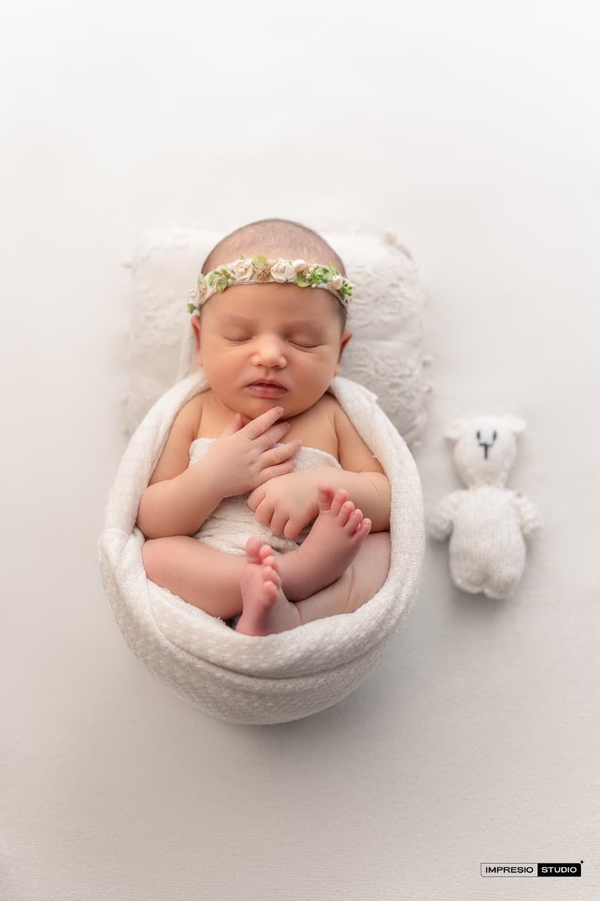 Newborn baby sleeping in white bowl with floral headband beside a small white teddy bear on white background.