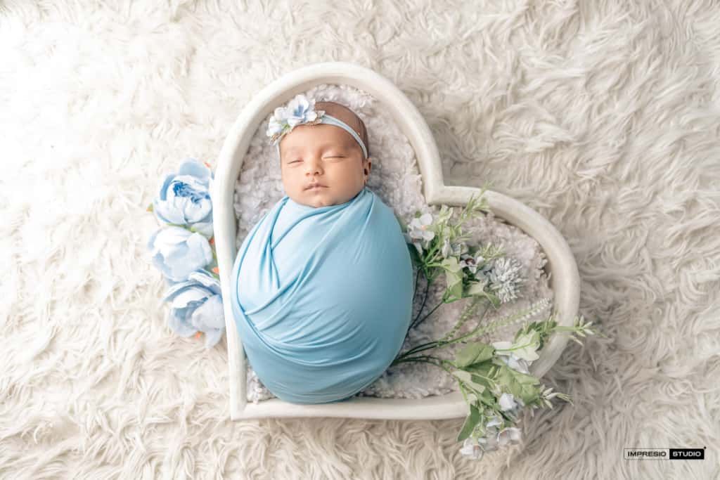Newborn baby swaddled in blue wrap sleeping in white heart-shaped prop with floral accents on fluffy white rug.