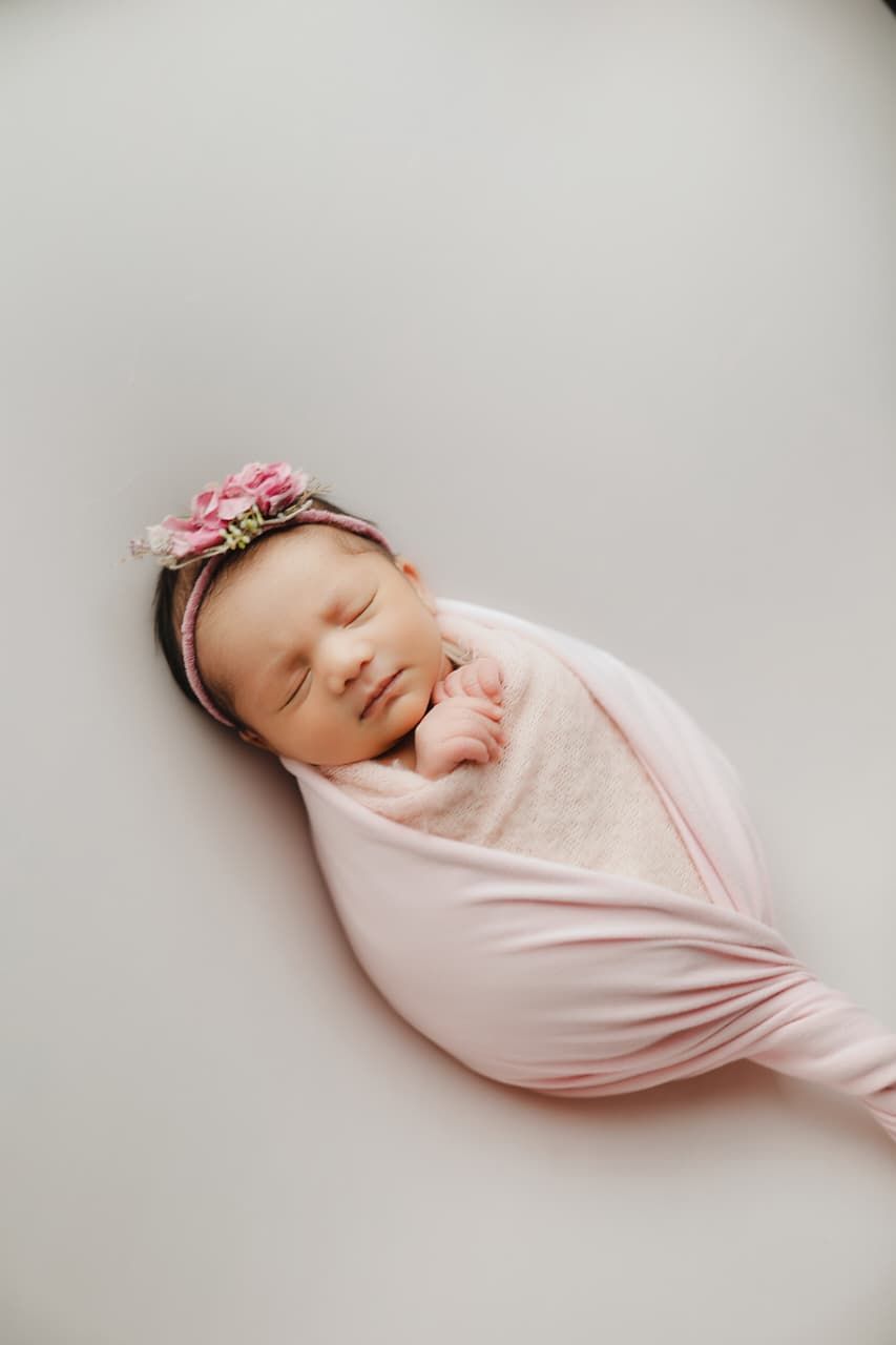Sleeping newborn baby swaddled in pink wrap with floral headband on white background.