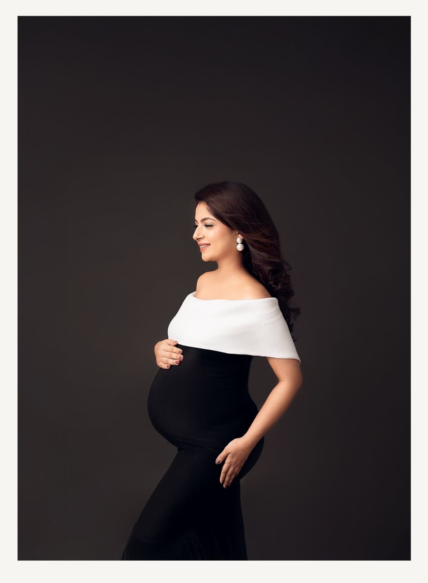 Black and white maternity photo with off-shoulder gown showing silhouette against dark backdrop.