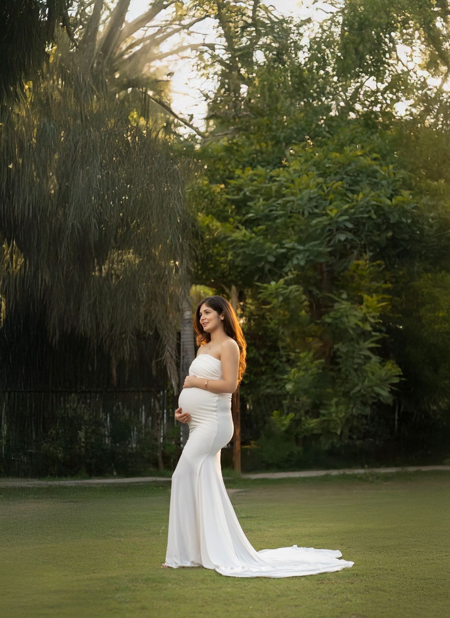 Elegant maternity photo on manicured lawn with sunlit trees in background.