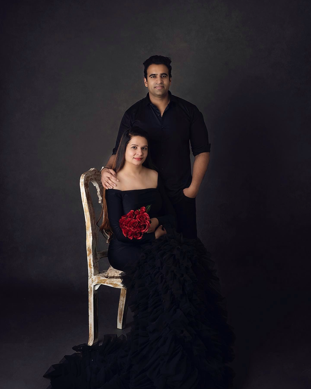 Elegant studio maternity photoshoot featuring a black dress and red roses on a dark background.