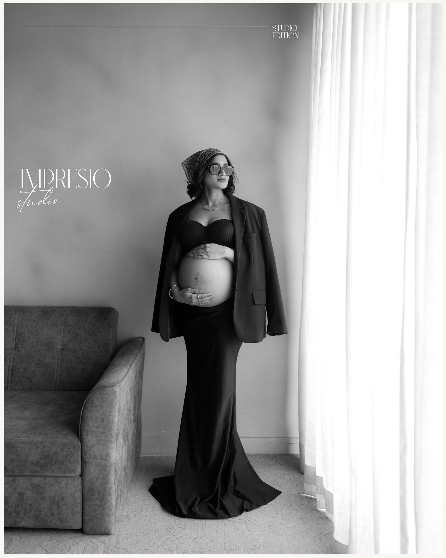 Elegant pregnant woman in black gown and blazer poses by window in stunning maternity portrait by Impresio Studio.