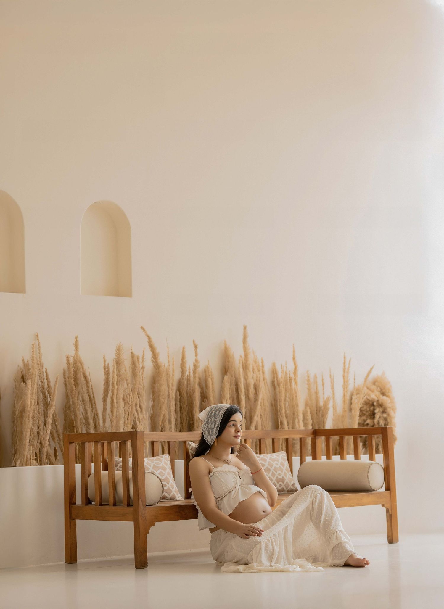 Pregnant woman in white dress sitting on wooden bench with pampas grass decor in a minimalist beige studio setting.