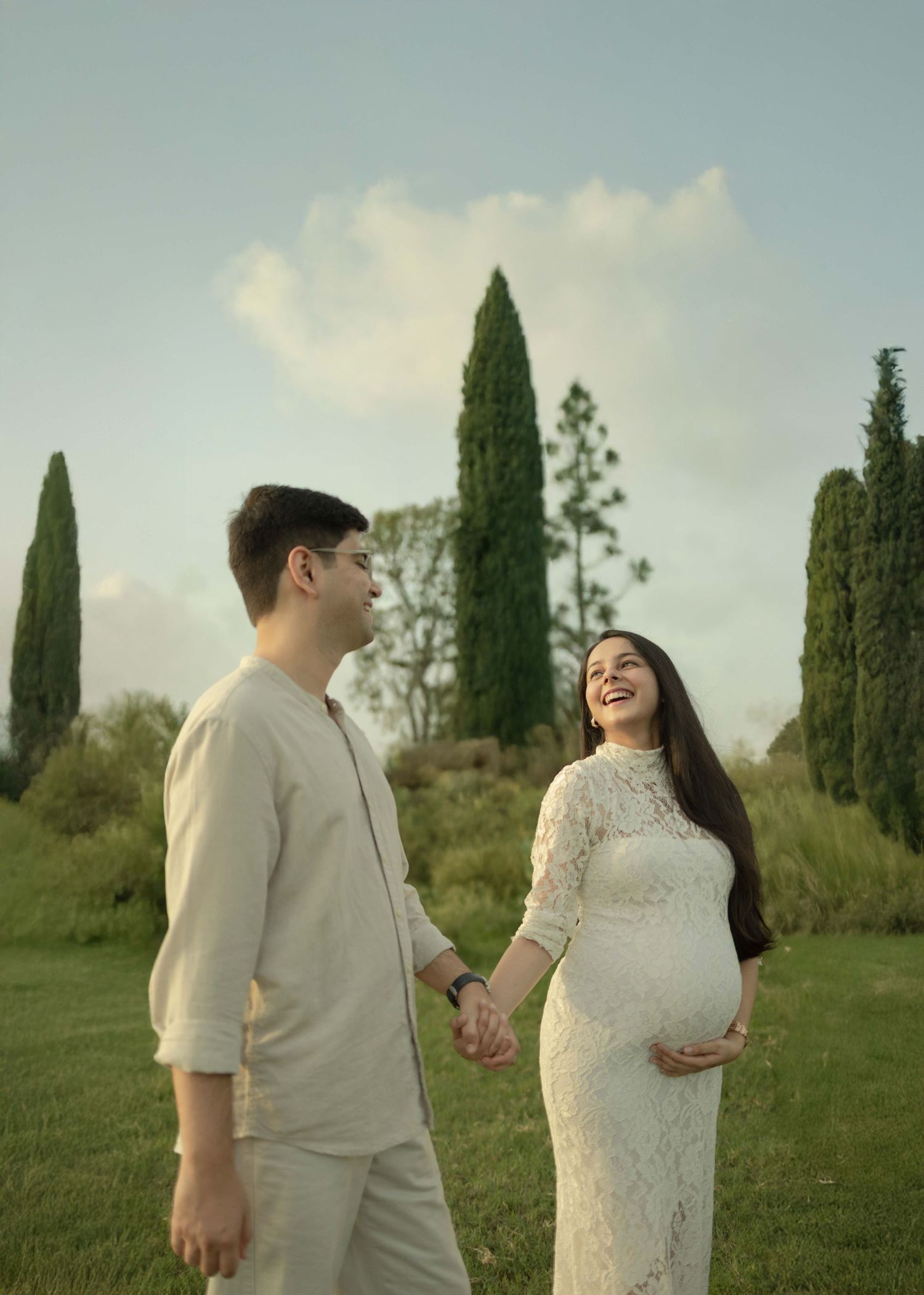 Pregnant couple holding hands in a garden, woman in white lace dress smiling, tall cypress trees in background.