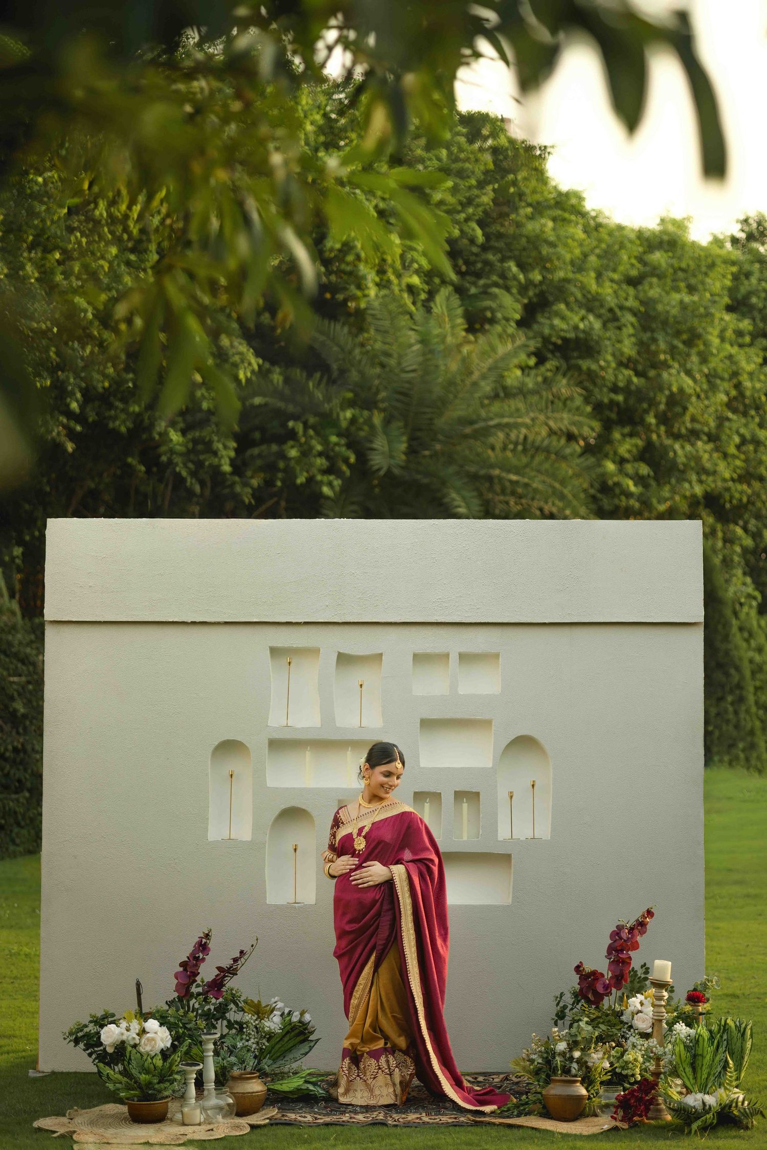 Woman in elegant maroon and gold saree poses before ornate white architectural backdrop surrounded by lush greenery and floral decor.