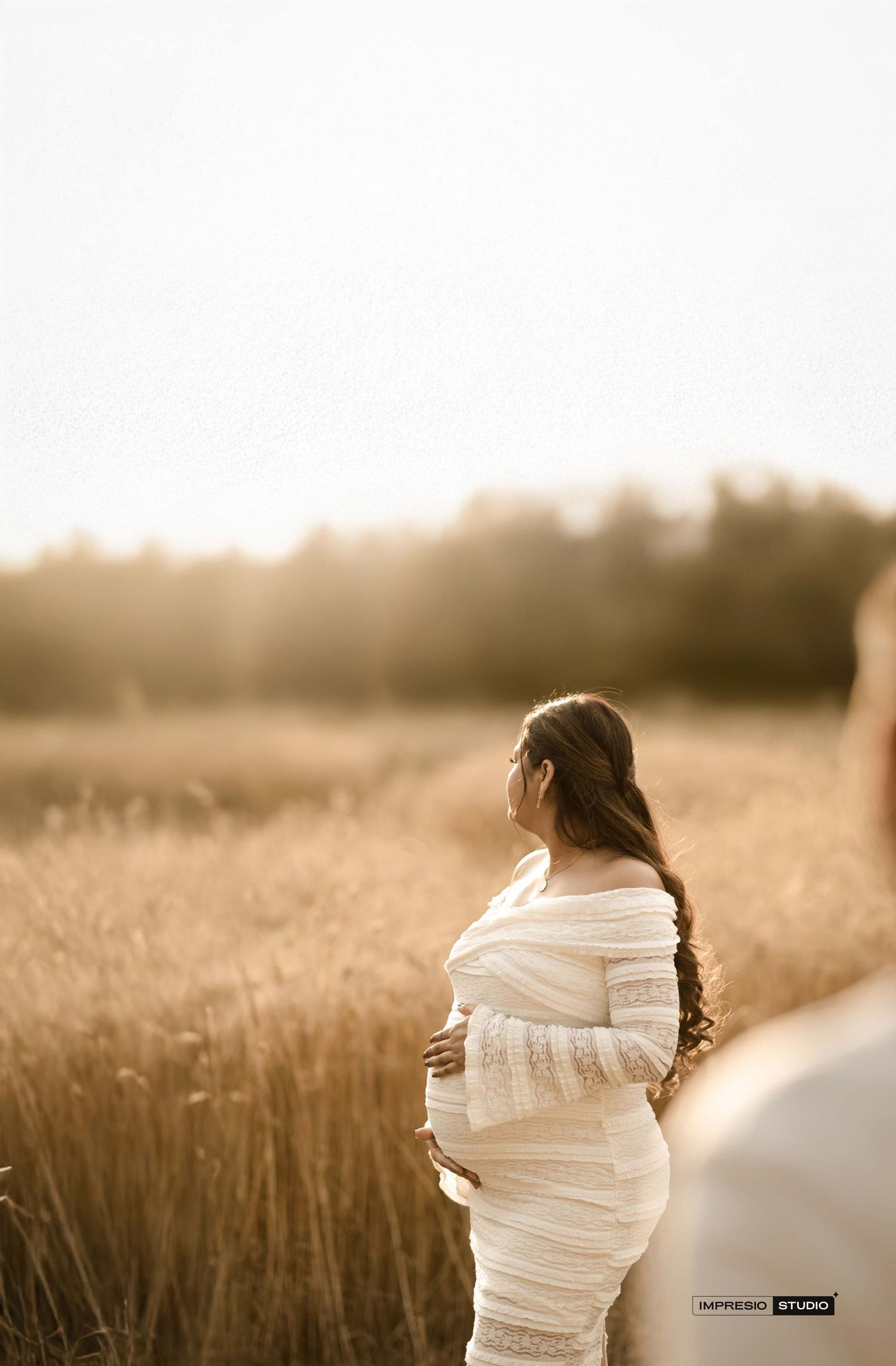 Pregnant woman in white lace dress standing in golden wheat field during sunset maternity photoshoot.