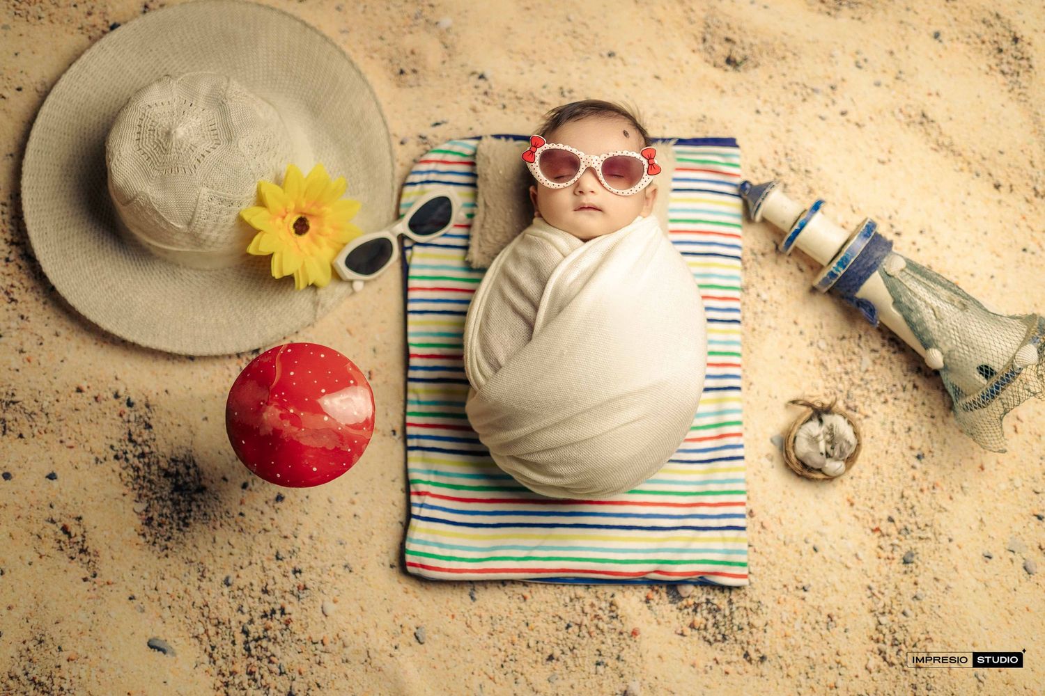Adorable newborn baby wrapped in white swaddle wearing pink sunglasses, posed on striped towel with beach accessories on sand.